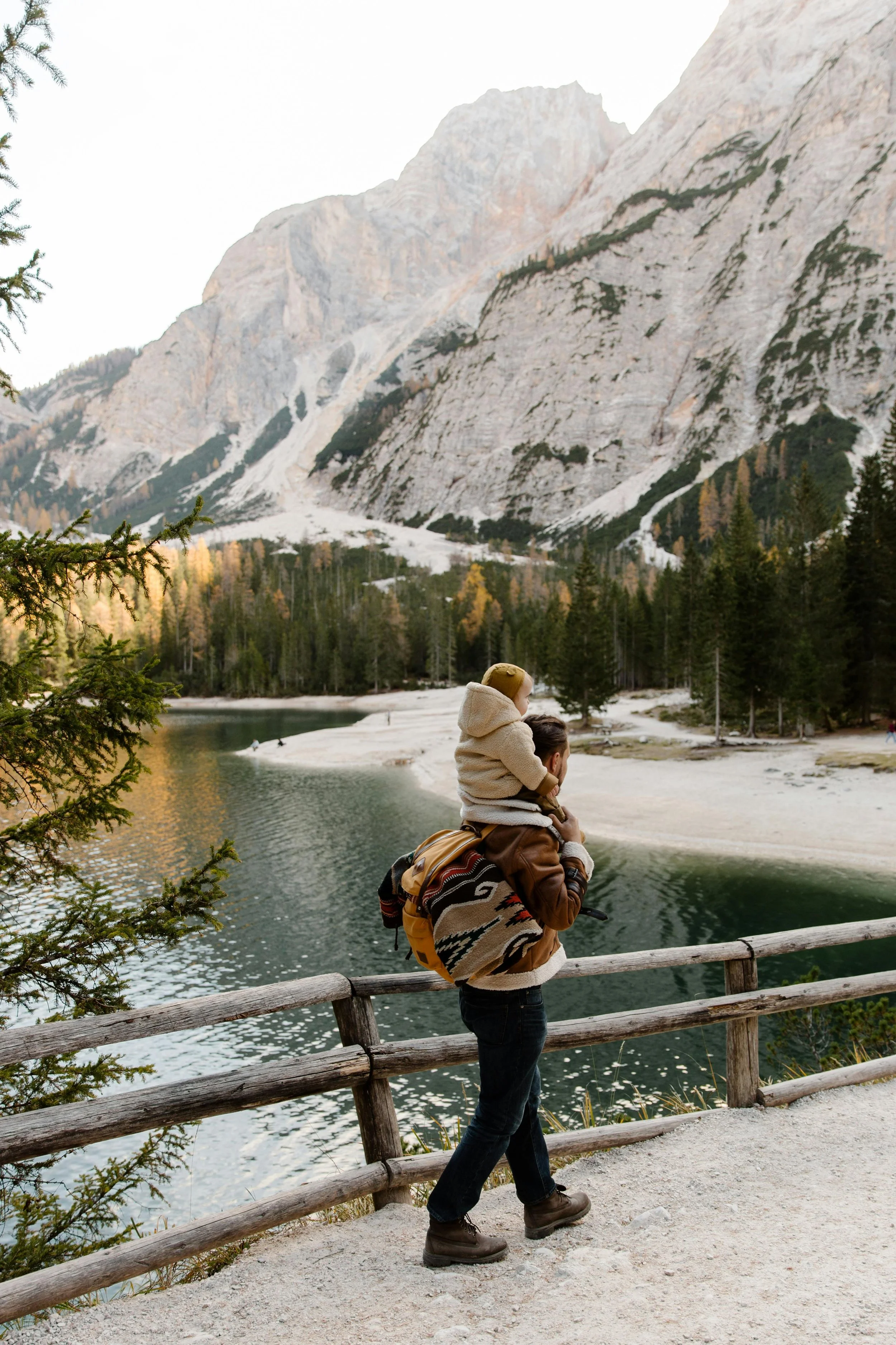 A person carrying a child on their shoulders by a lake with mountains and pine trees in the background.