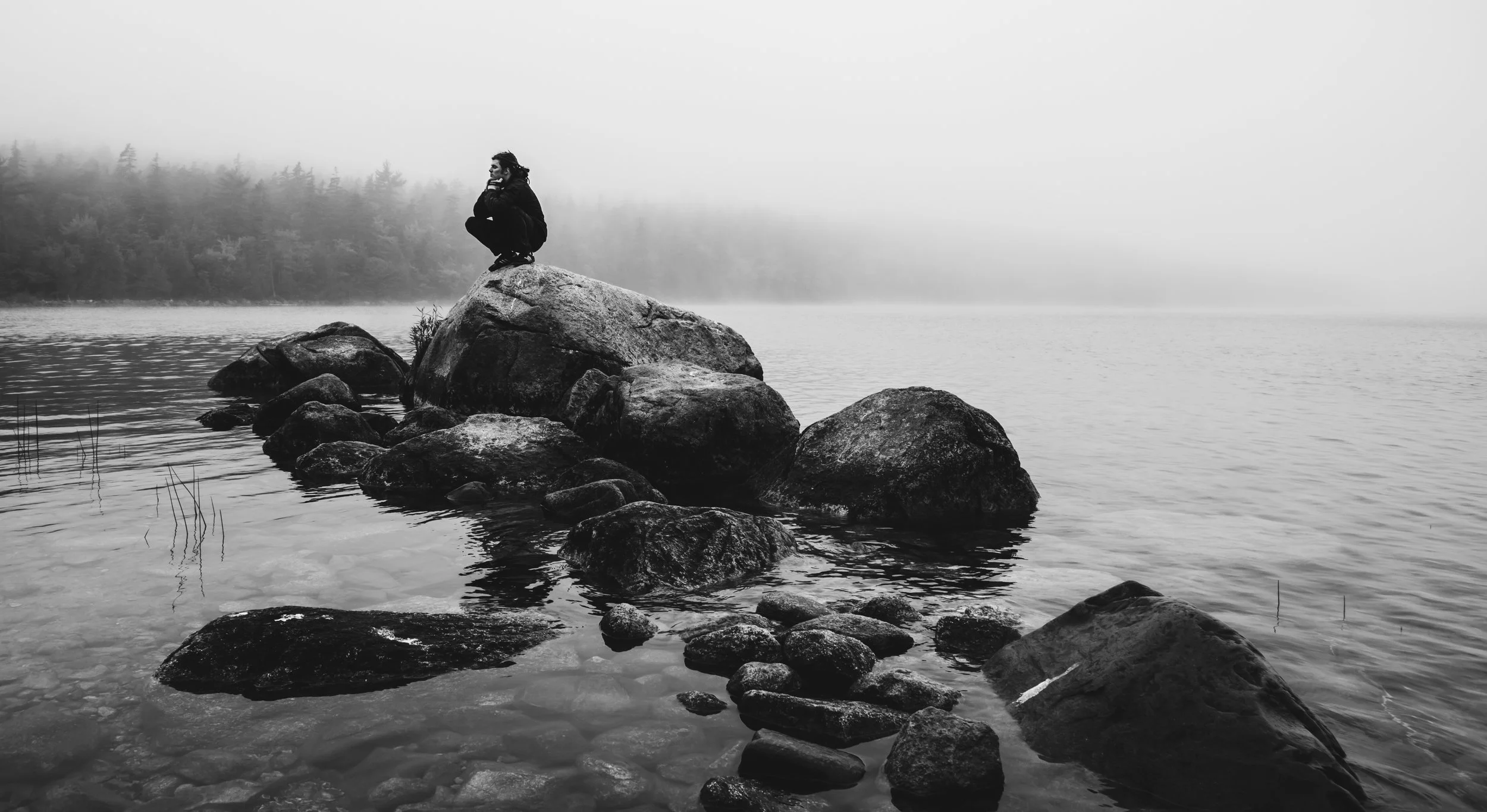 A person crouching on a large rock in a foggy lake with trees in the background, black and white photo.
