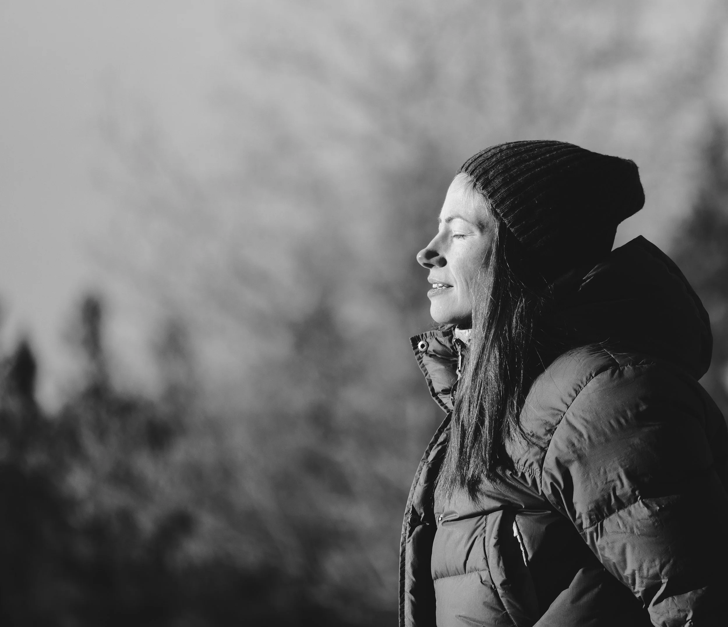 A woman with long hair wearing a knit hat and puffy jacket, standing outdoors with eyes closed and a peaceful expression.
