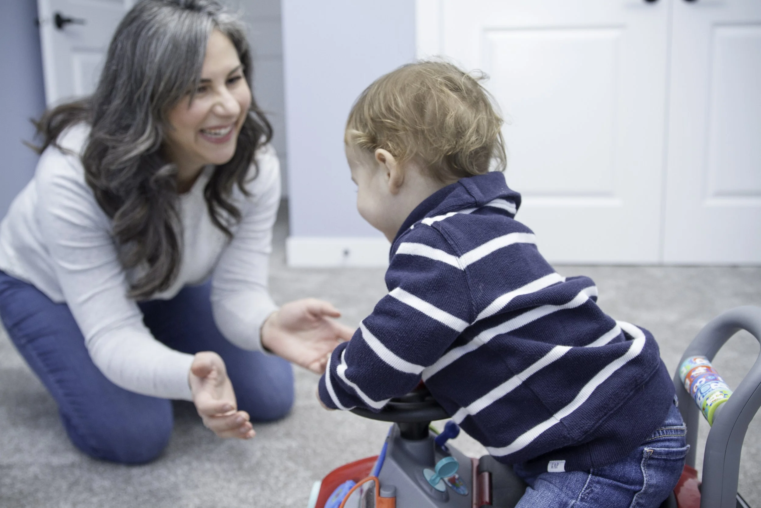 A woman kneeling down and smiling at a young boy with red hair, who is sitting on a tricycle. They are indoors in a room with white doors and gray carpet.