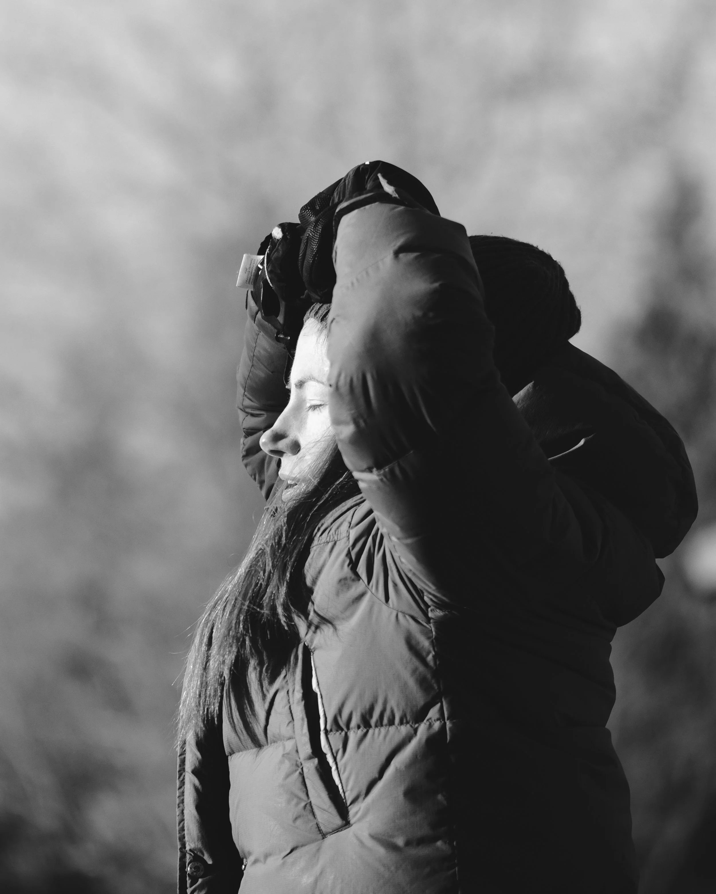 A person wearing a hooded jacket, carrying a backpack, with long hair, and his or her face in profile, under a cloudy sky.