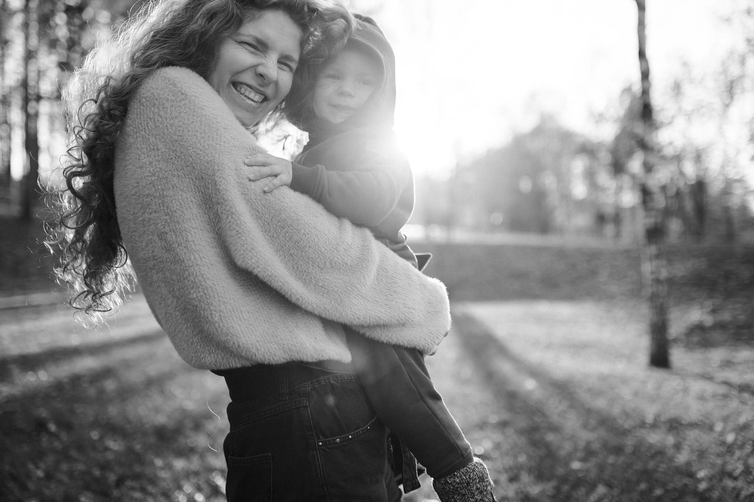 A woman with curly hair smiling and hugging a child in an outdoor park with trees, illuminated by sunlight in black and white.