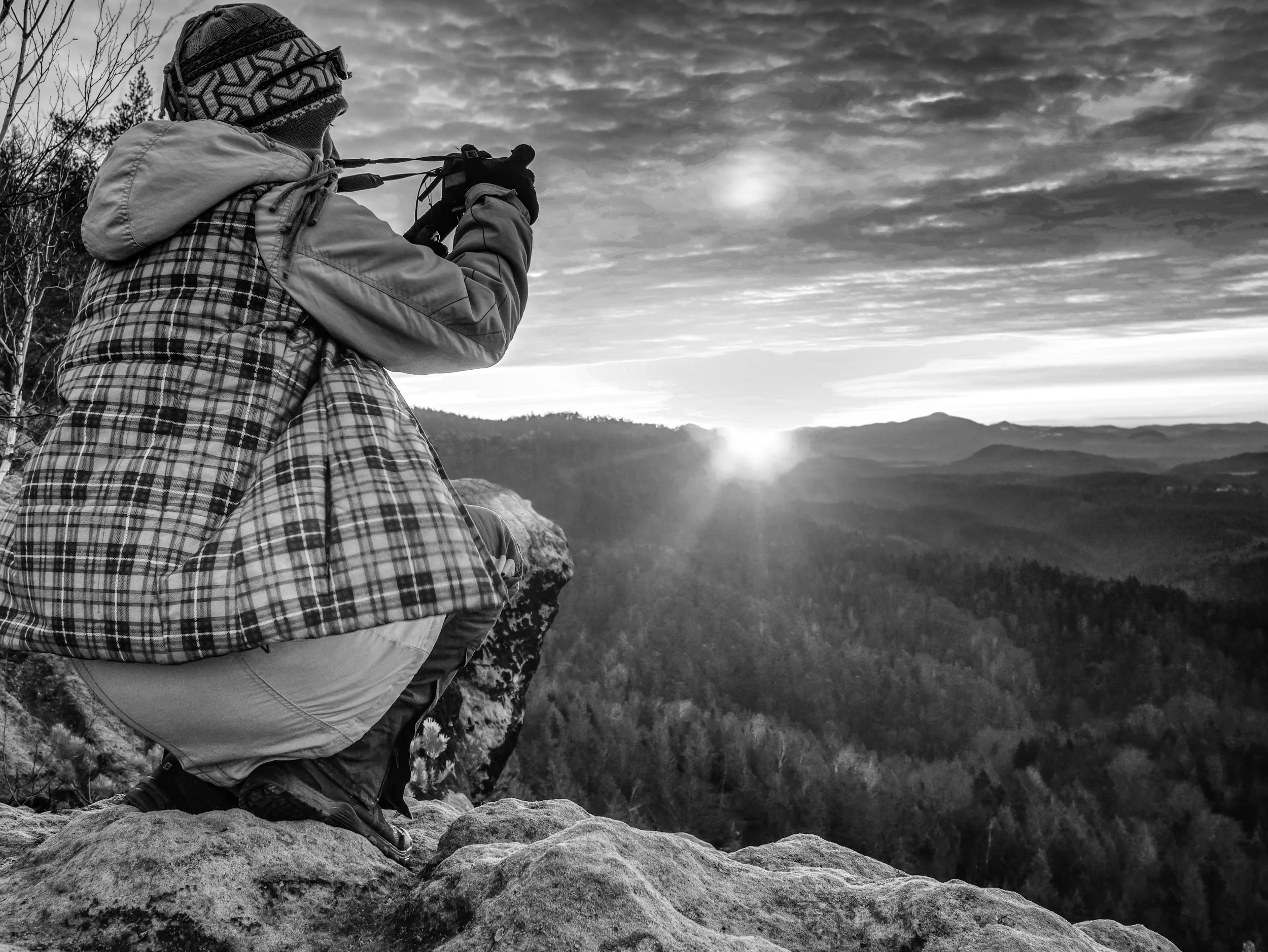 Person sitting on rocks taking a photo at sunrise or sunset over a mountainous landscape in black and white.