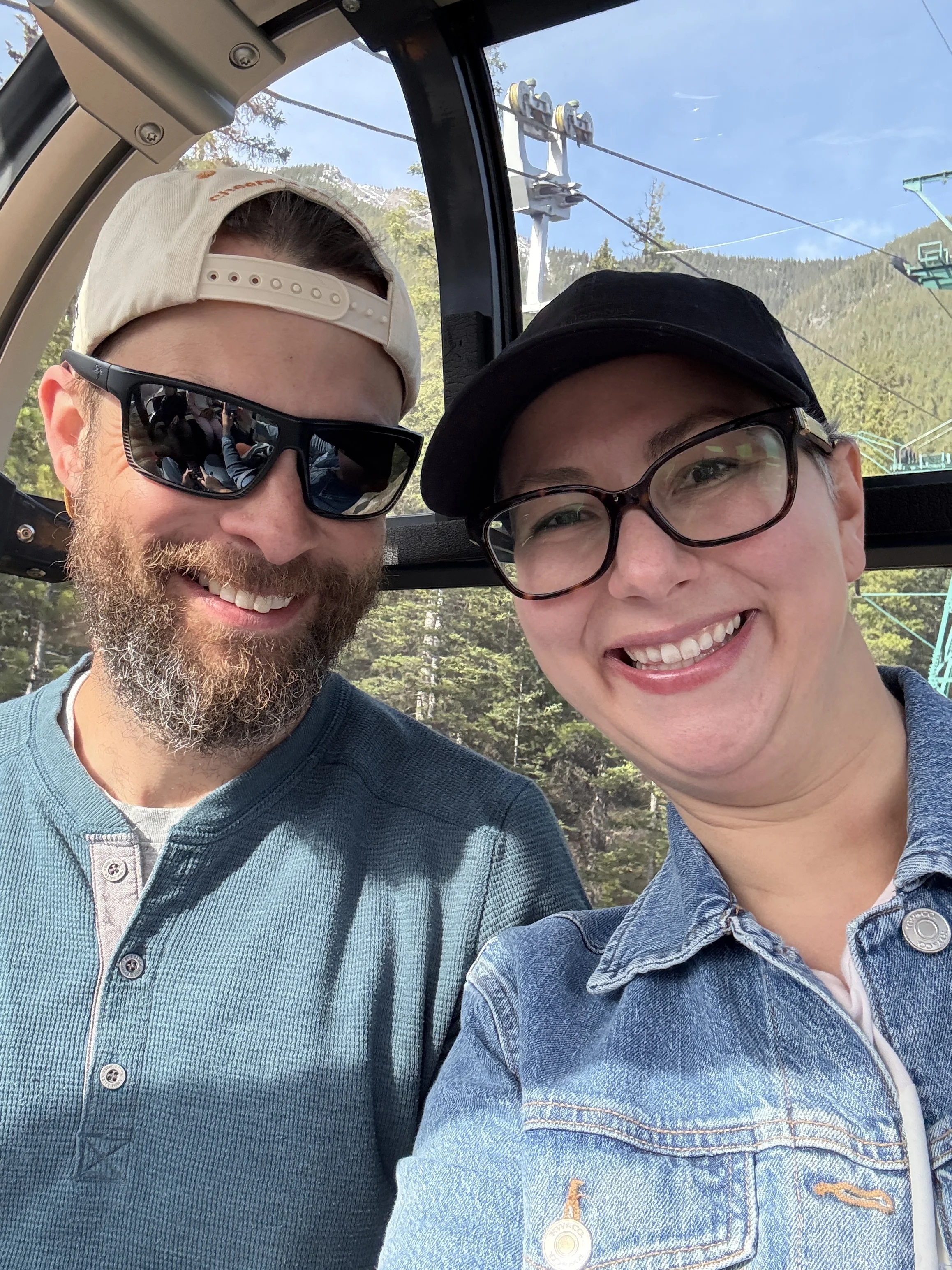 Two smiling people taking a selfie inside a ski lift with a mountain and trees in the background.