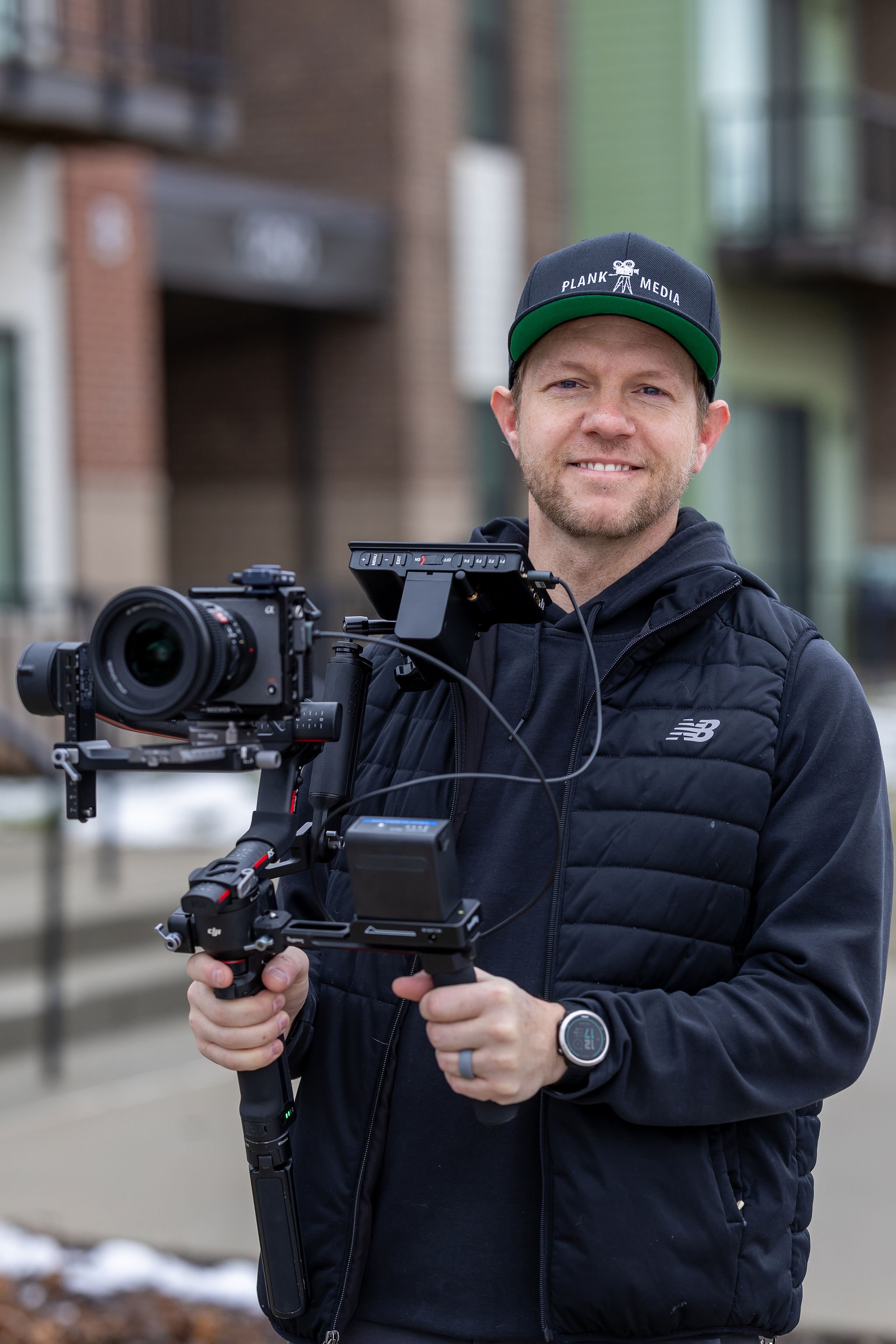 A man with a beard and short hair wearing a black jacket and a watch, holding a professional camera stabilizer with a camera mounted, outdoors in front of buildings with a blurred background.