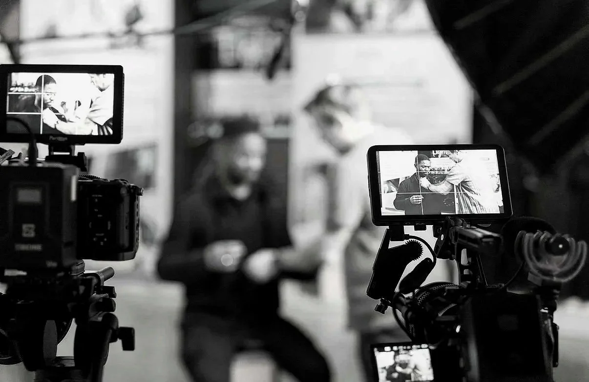 Black and white photo of a camera filming a man in a hoodie, with a person in a striped shirt adjusting his collar, in an urban setting.
