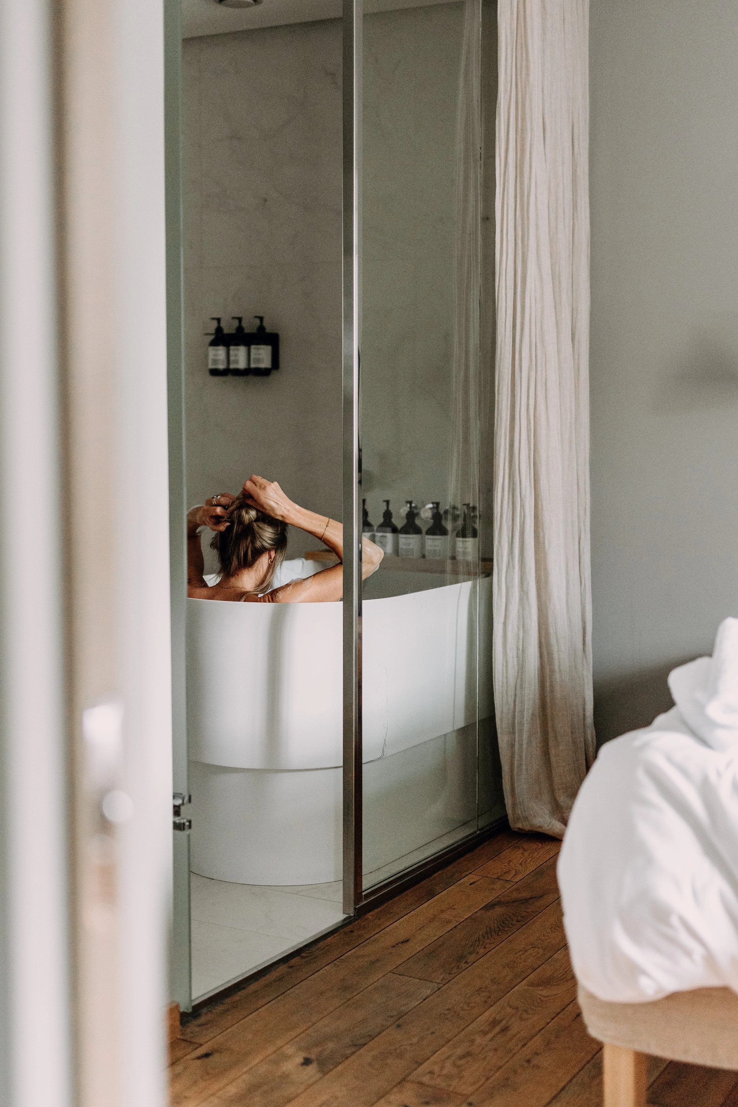 Person som vasker håret i badekaret, sett gjennom et speil og en glassdør i badet.