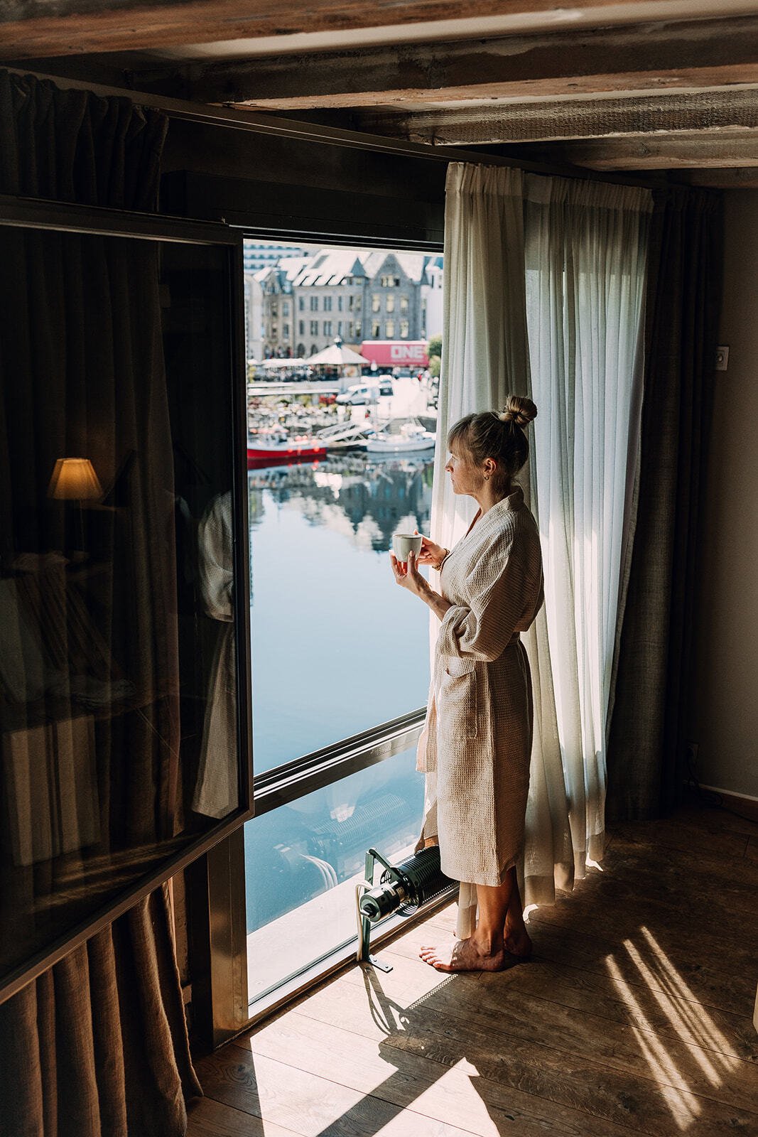 En kvinne i morgenkåpe står ved et vindu og drikker kaffe, med en marina og bygninger i bakgrunnen.