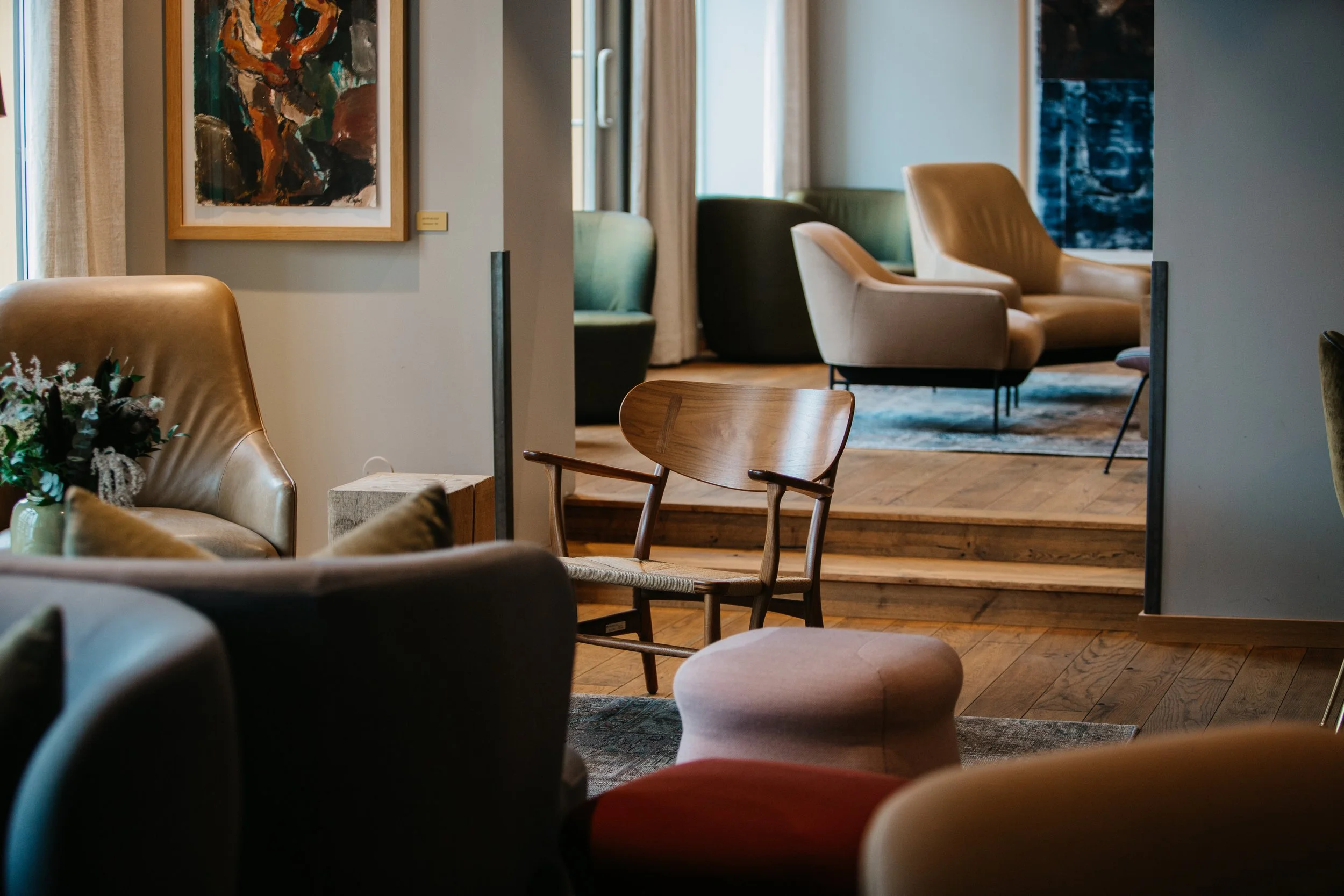 Innendørs rom med flere stoler i ulik farge og design, veggkunst, gulv og tepper, og store vinduer med gardiner