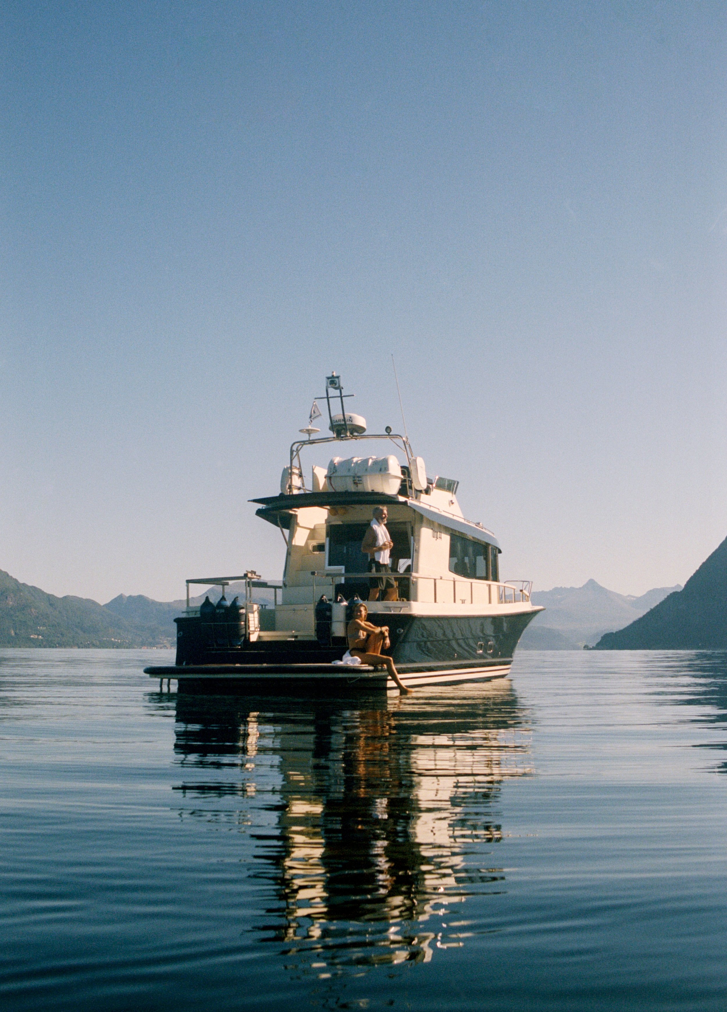 En yacht som ligger rolig på vannet med fjell i bakgrunnen. To personer er synlige på yttersiden av båten, og båten reflekteres i vannet.