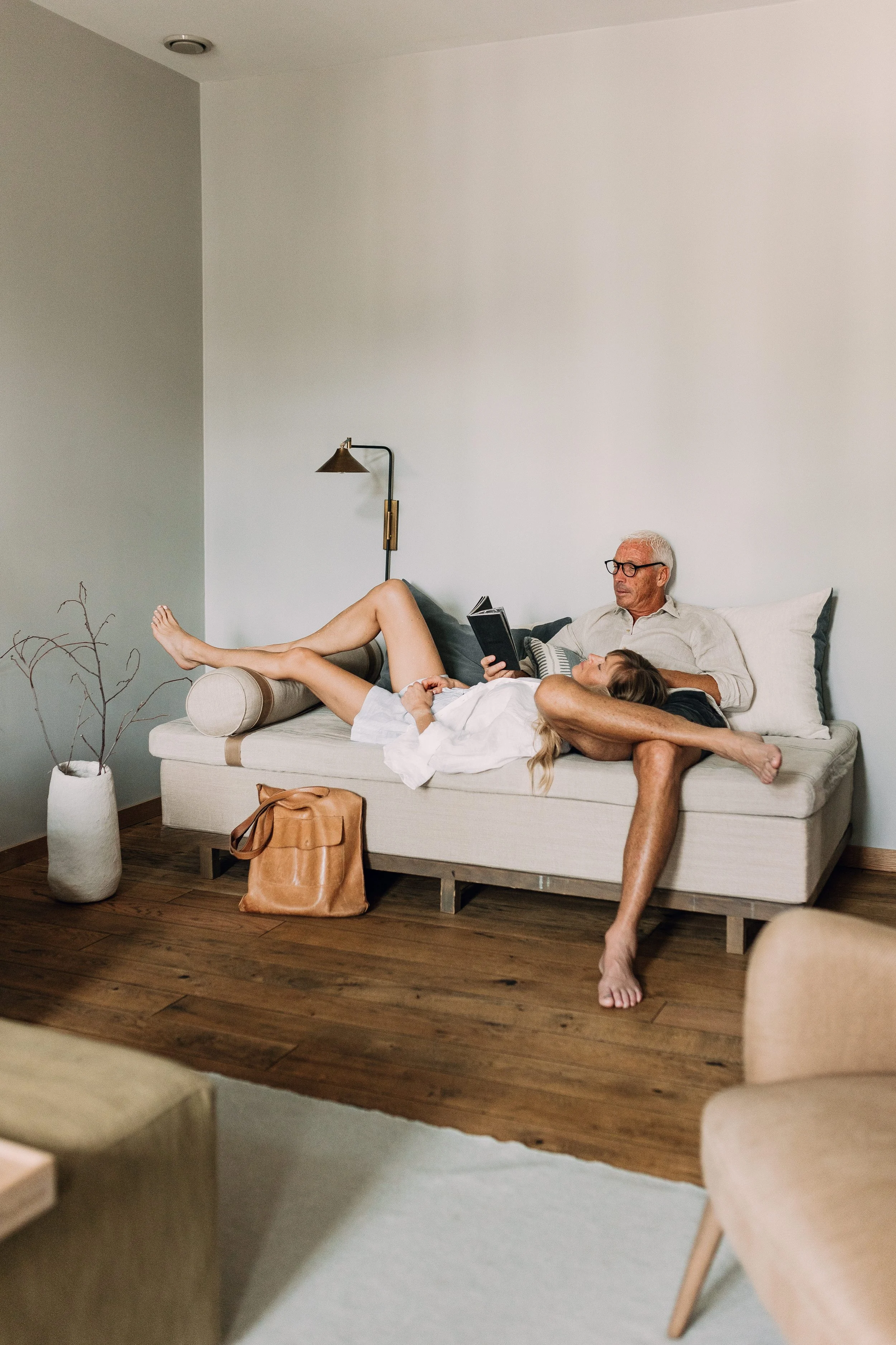 Et rom med en sofa der en eldre mann med hvitt hår og briller leser en bok, mens en yngre kvinne med langt hår ligger med hodet i fanget hans. Kvinnen har bena opp på sofaen, og det er en plante i en lys keramikkvasspott til venstre.