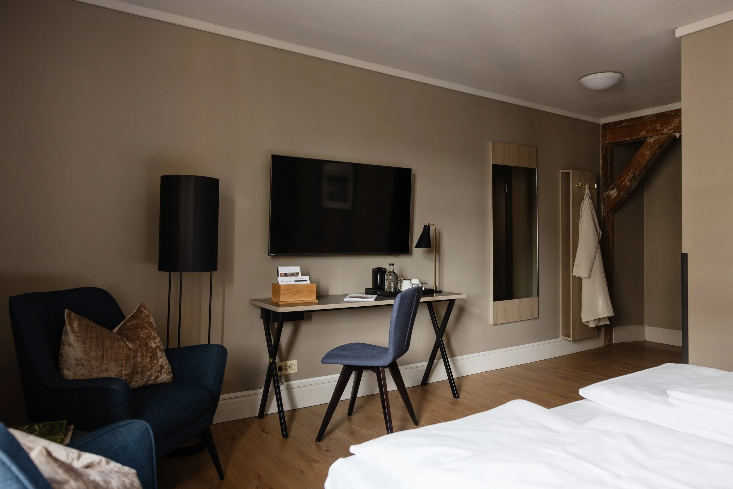 Innendørs rom med veggmontert TV, skrivebord med lampe og stoler, sofa med pute, og en åpen garderobe med klær.