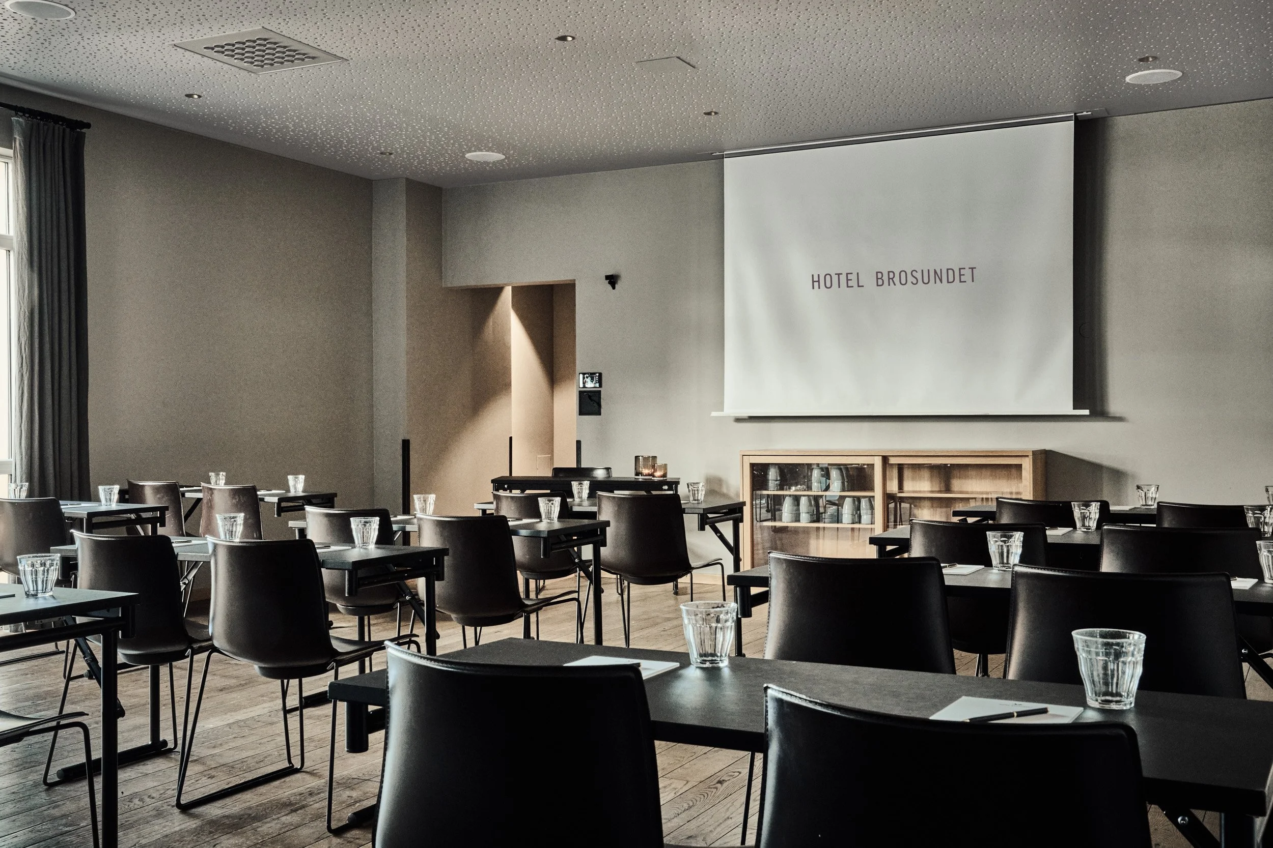 Et møterom med flere bord og stoler, en stor projeksjonsskjerm med teksten 'HOTEL BROSUNDET', vinduer med gardiner, og glass på bordene.