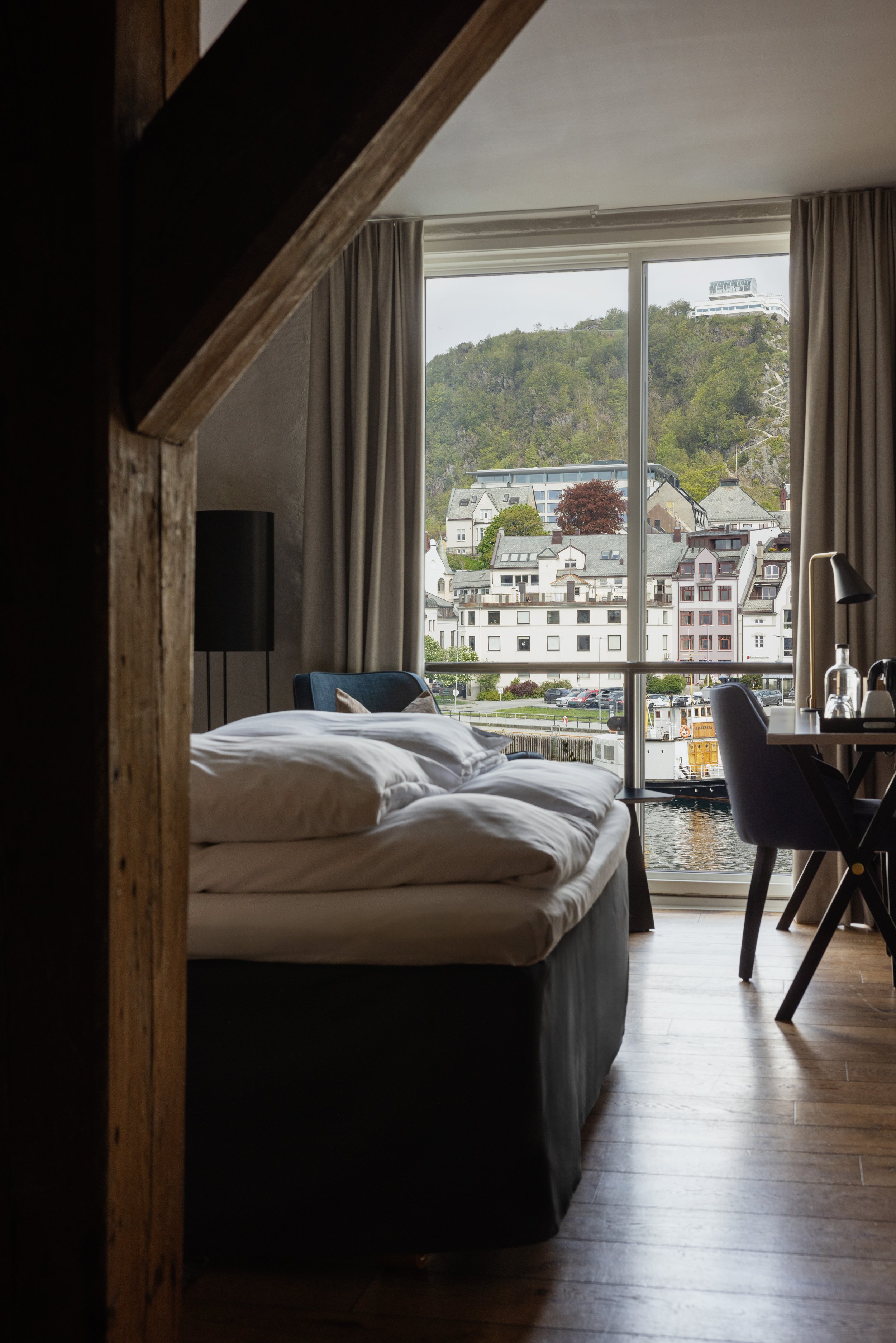 Innside av et rom med seng, vindu som viser en by med fjell og bygninger, og et arbeidsbord med stol.