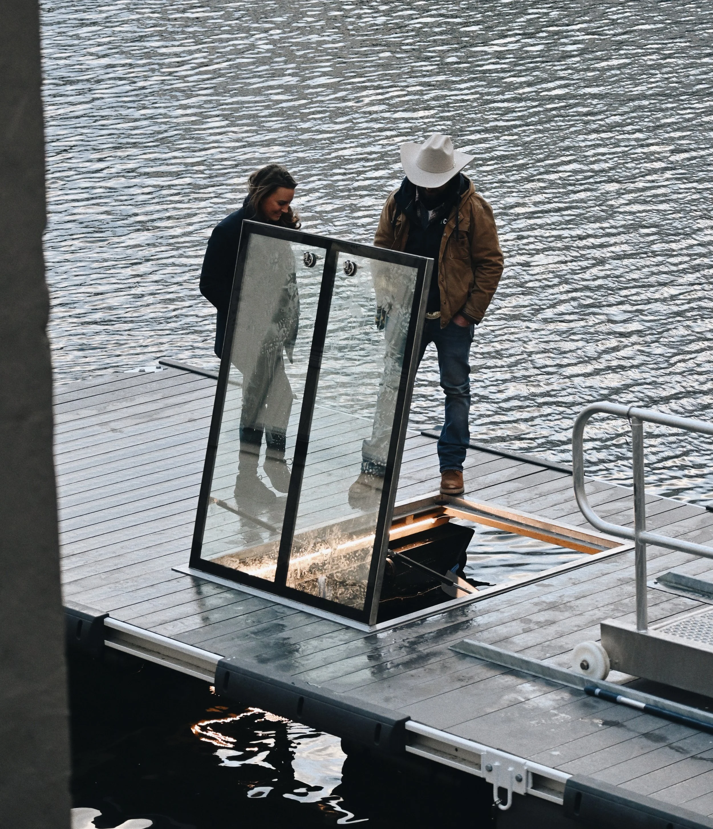 To personer ser på en speil som er satt opp på en kai ved vannet. En av dem har på seg stor hvit hatt, og den andre har langt hår og mørk jakke.