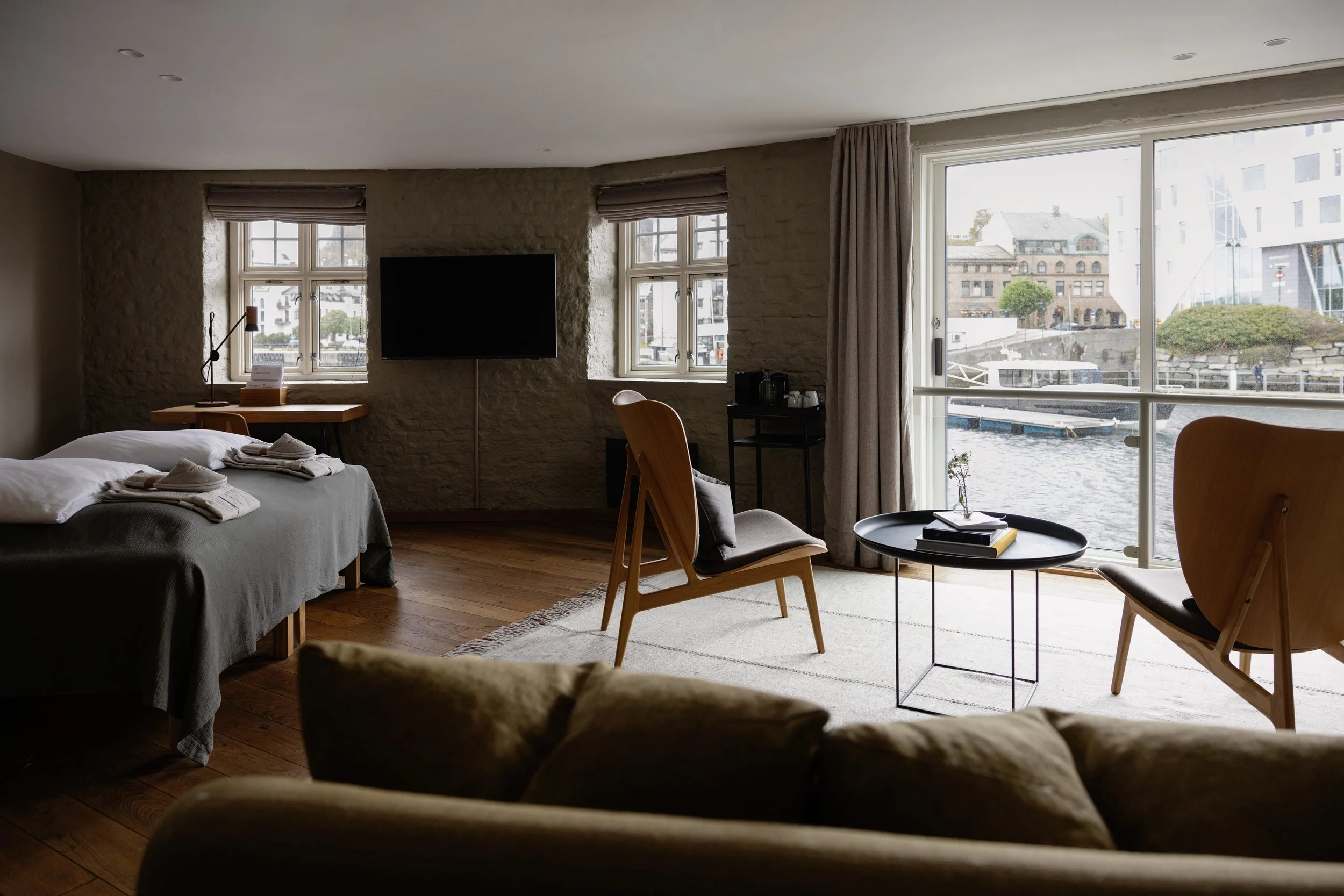 Et hotellrom med to senger, en stol, et lite bord med bøker, et vindu med utsikt over vann og bygninger, og en TV på veggen.