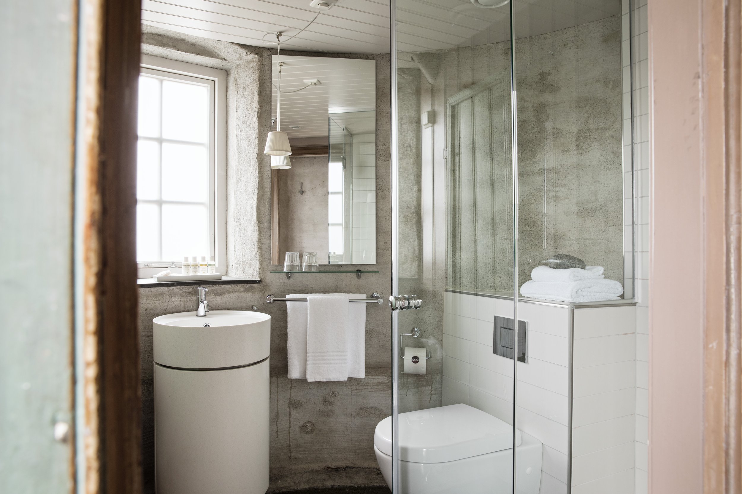 Utsikt inn i et moderne bad, sett gjennom en dør med rustfritt stålramme. Badet har en hvit toalett og en sirkulær vask på veggen. Det er to glassdører som skiller dusjområdet med en innsats av hvite fliser. På vinduskarmen er det små krus og personlige eiendeler.