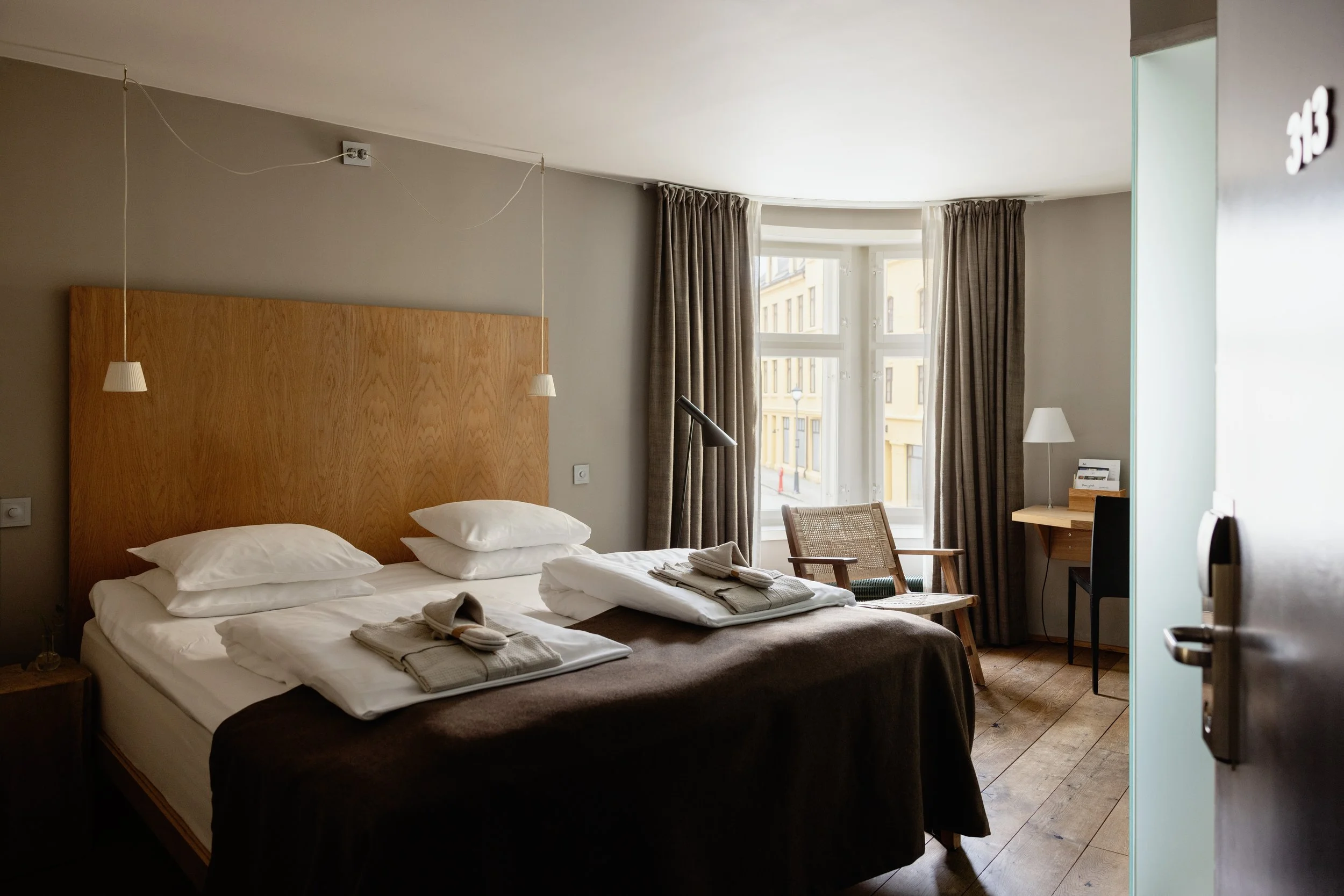 Et hotellrom med to senger, vindu med gardiner, skrivebord, stol, bordlampe og et nattbord med lamper.