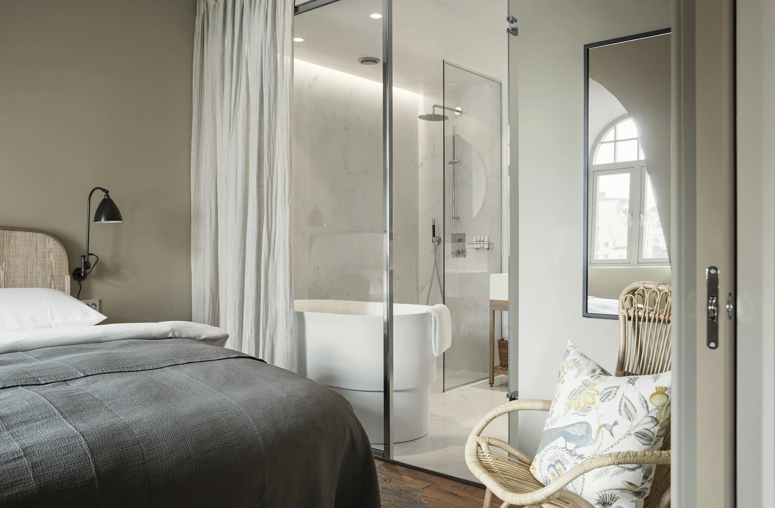 Soverom med seng, svart nattbordslampe, og bad med dusj og badekar sett gjennom en glassvegg.