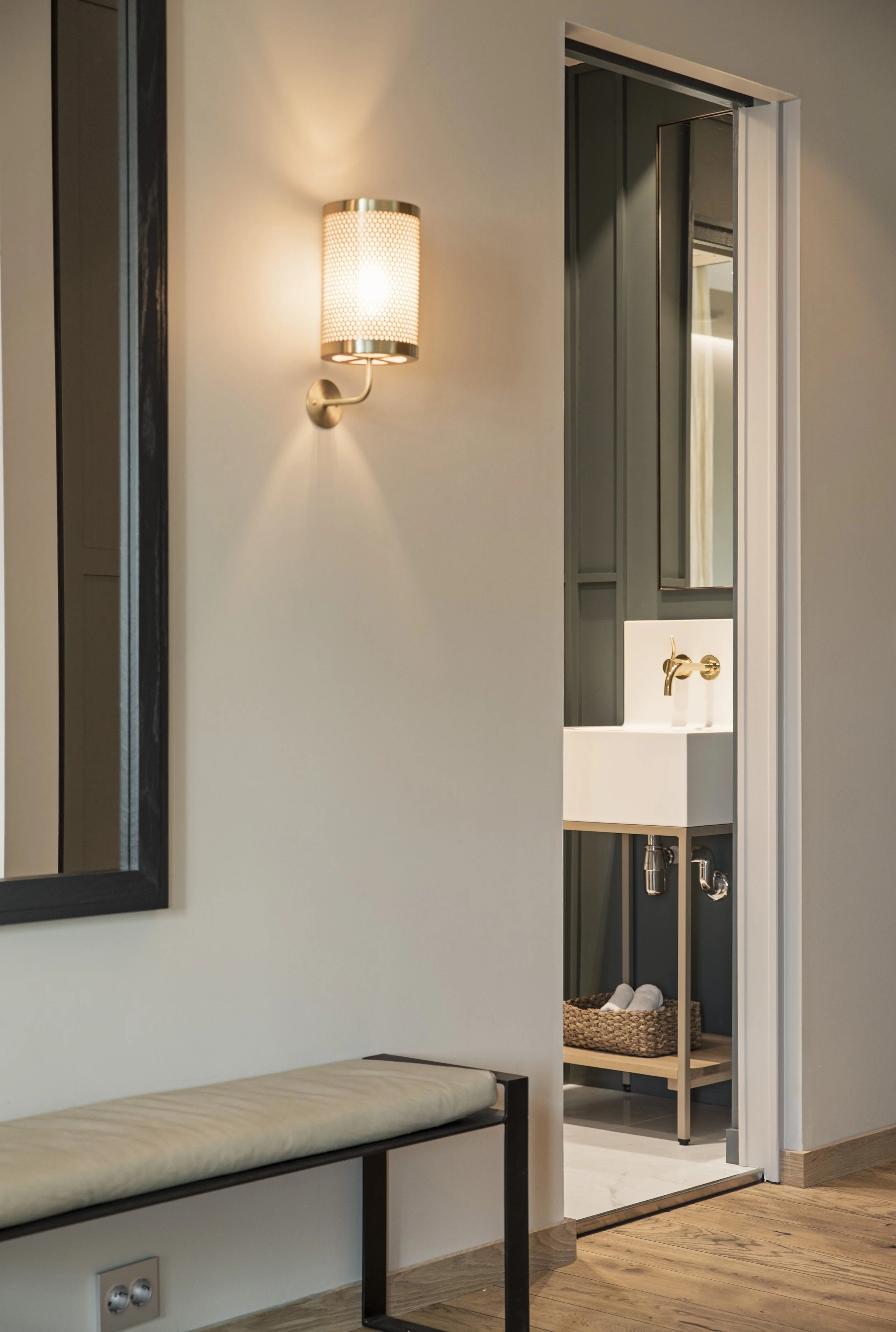 Veggen er utstyrt med en vegglampe som har et mønster av små hull. Det er en speilvegg på høyre side av bildet, og bak den speilet er et rom med en liten vask, en kurv med håndklær og andre baderomsartikler.