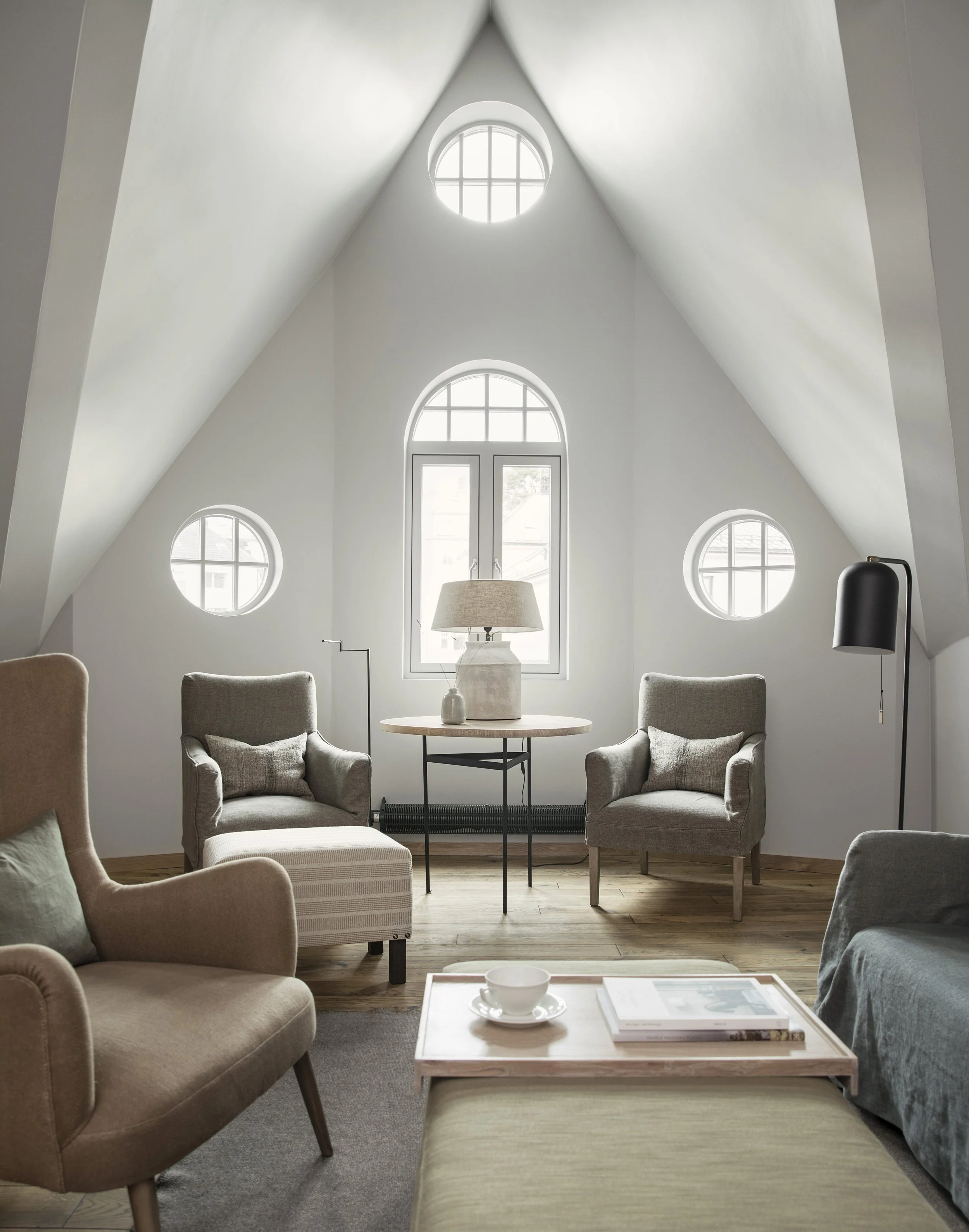 Et lyst rom med hvite vegger og mange vinduer. Det er flere stoler, en mørk vifte, en lampe og en tabell med en kopp og en bok.