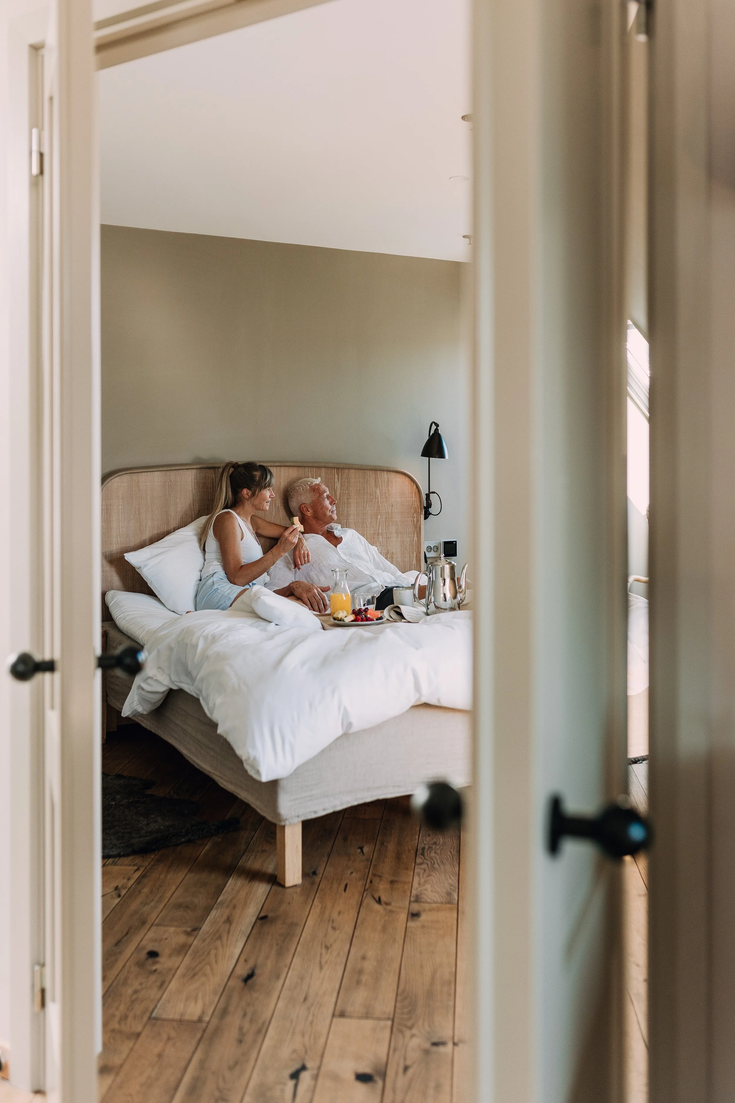 To personer ligger i en seng på et hotellrom, sett gjennom en åpen dør. Den ene personen er eldre med grått hår, den andre er yngre med mørkt hår i en hestehale, de smiler og har et glass juice og noe mat på nattbordet.