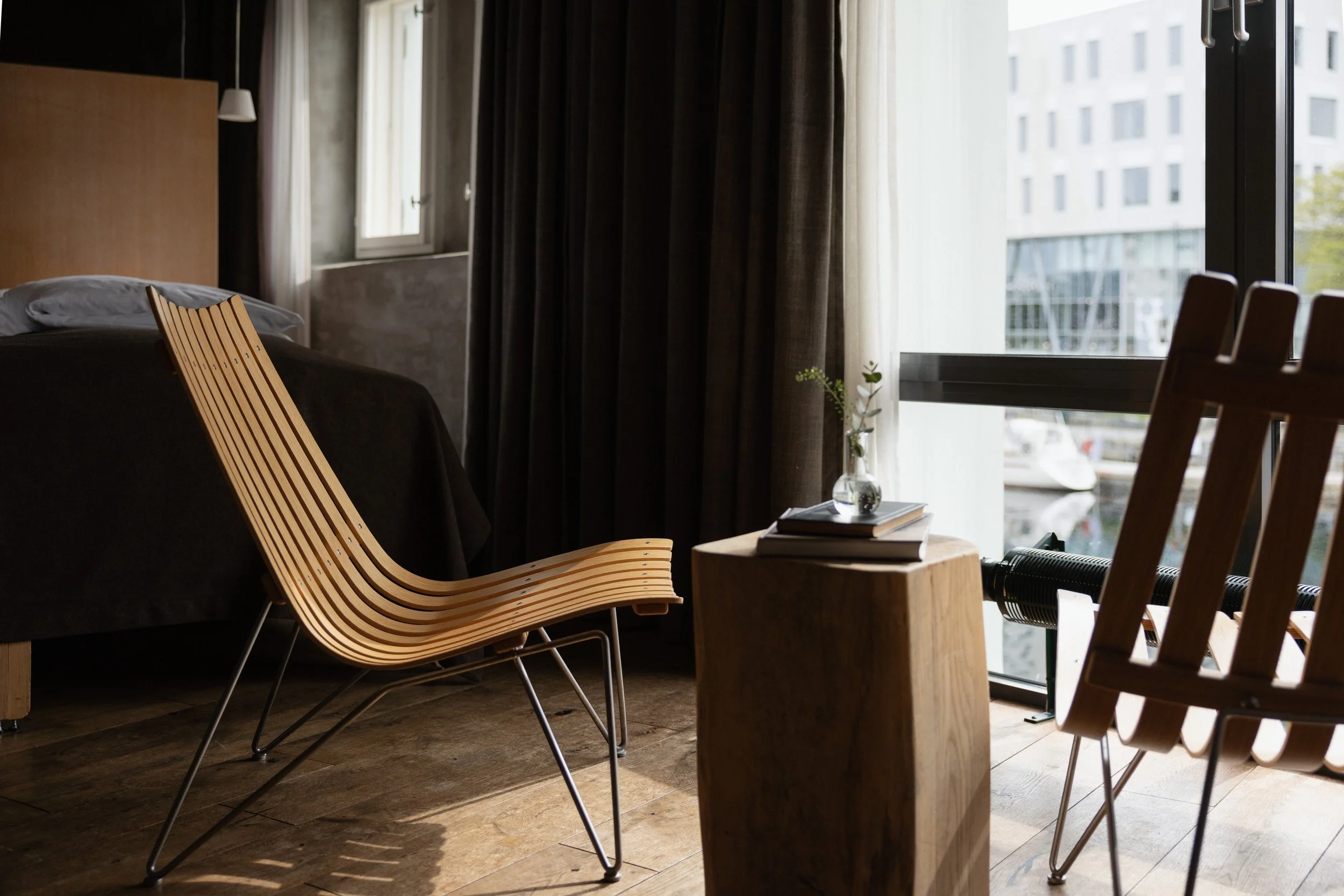 Innendørs rom med store vinduer, trestoler, en seng med svarte laken, og et bord med bøker og en glassvase med grønt plantestem. Utsikt over moderne bygninger utenfor.