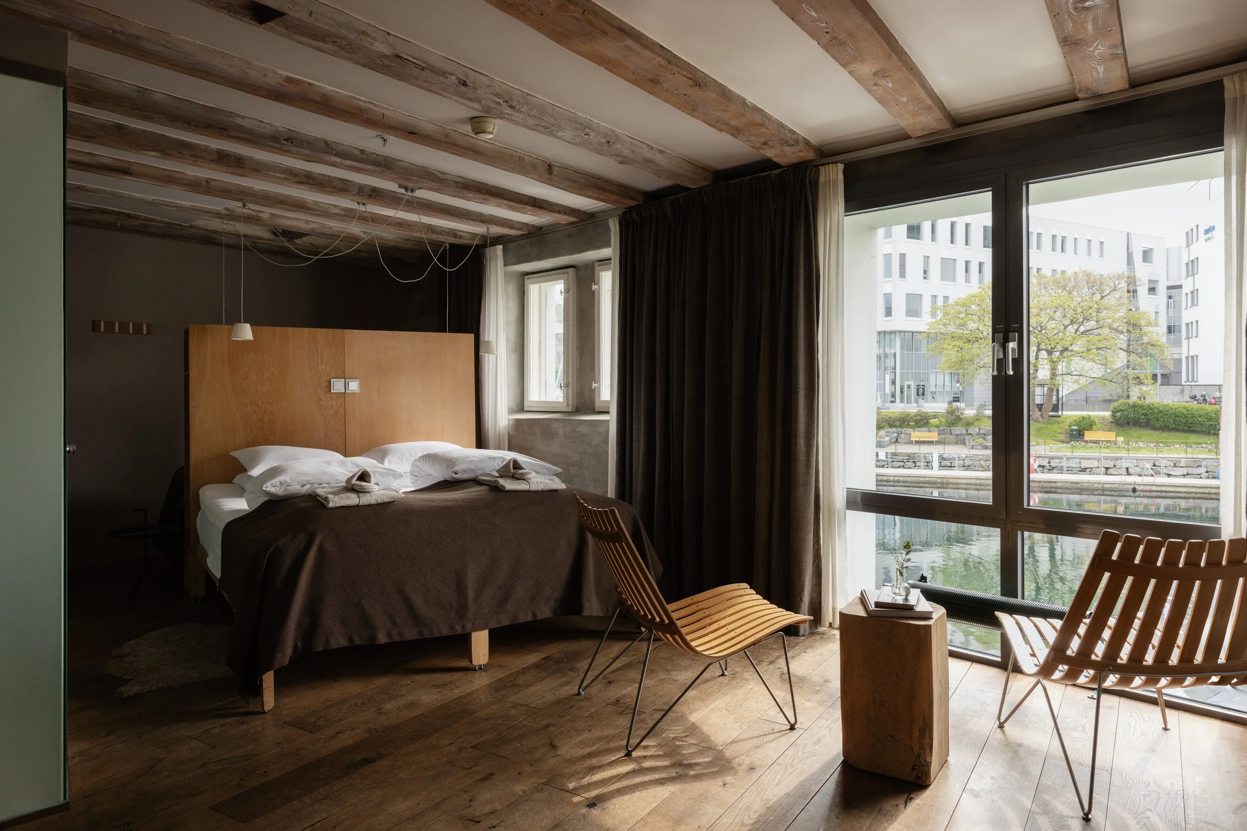 Innendørsrom med seng, store vinduer, trestøttede takbjelker, og moderne bygårder utenfor.