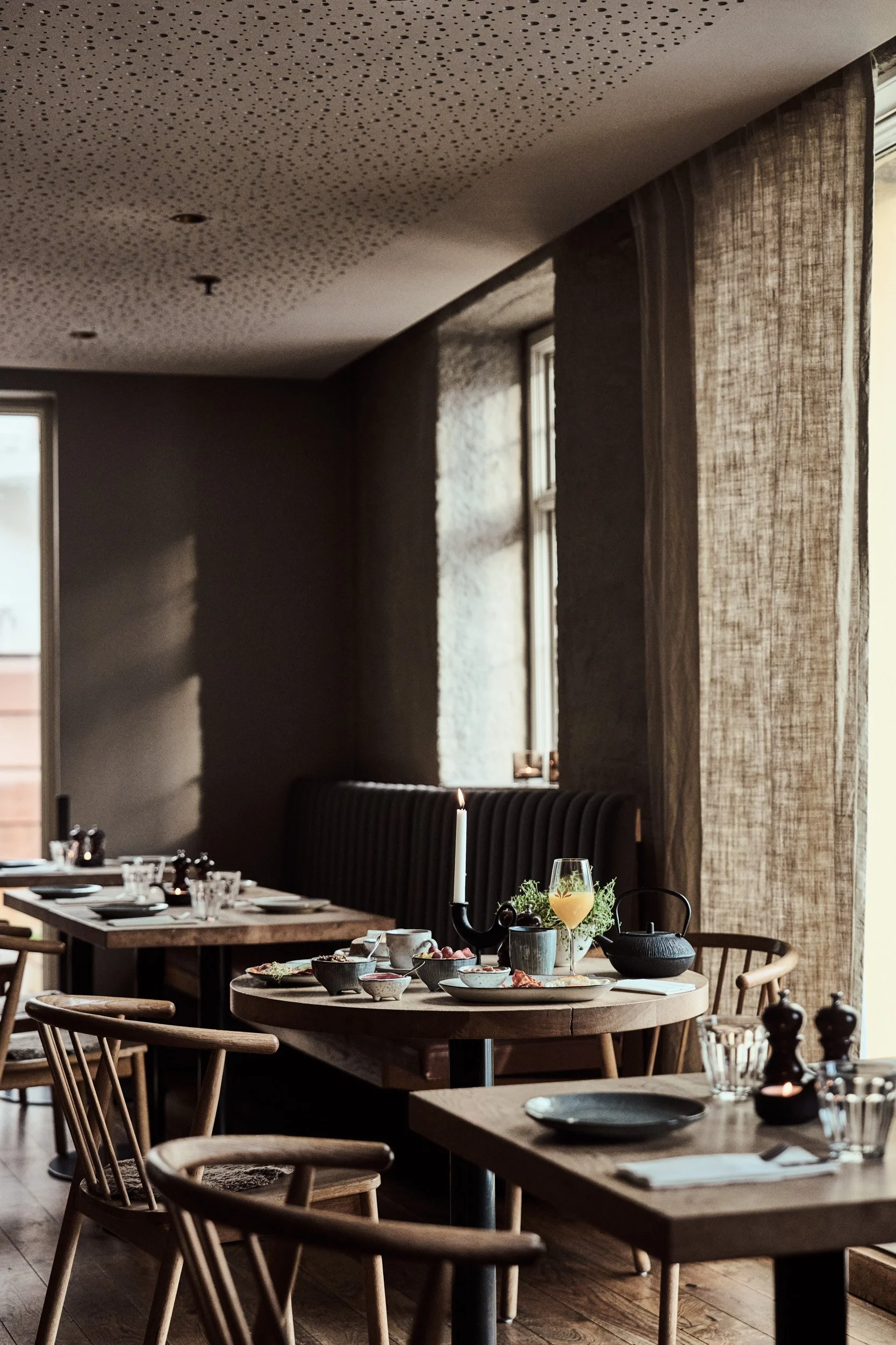 Et koselig spiseområde med oppdekkede bord og telys i en restaurant eller et hjem. Bordet har servise, glass og blomsterdekorasjoner. Det er store vinduer med grove, lyse gardiner som slipper inn naturlig lys.