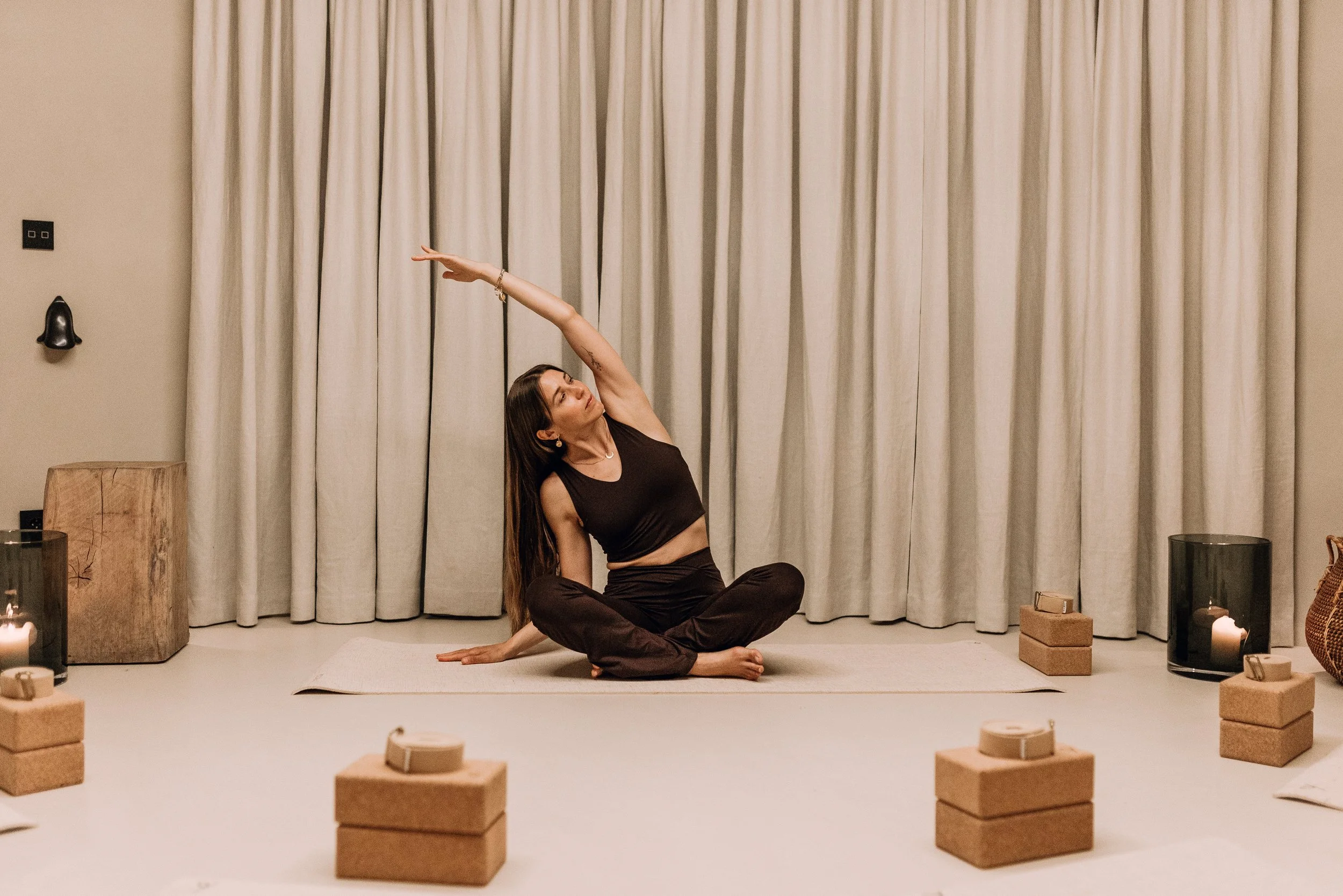 En kvinne som trener yoga eller meditasjon sittende på gulvet i et rom med beige gardiner. Røde mursteinblokker og stearinlys er synlige rundt henne.