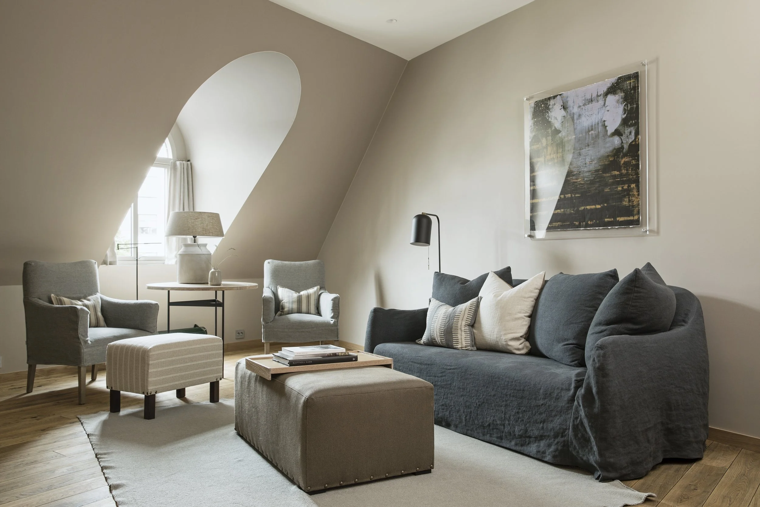 Lys, moderne stue med beige vegg, stor sofa med puter, to lenestoler, et trebord med bøker, et gulvlampe og kunst på veggen.