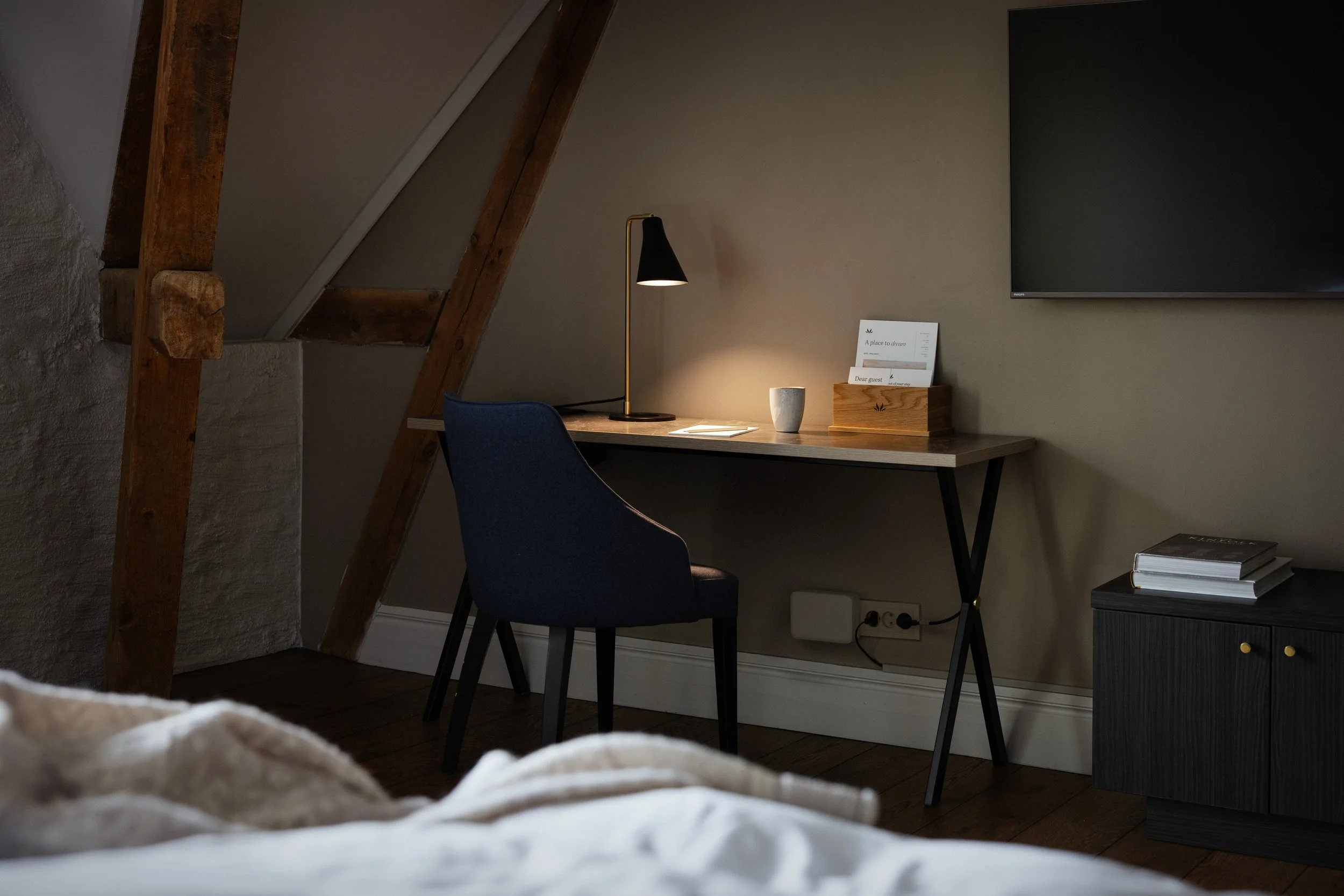 Et hjemmeslosekontor med et skrivebord, en svart stol, en bordlampe, kopper, bøker og et TV på veggen i et rom med skråtak og synlige trebjelker.
