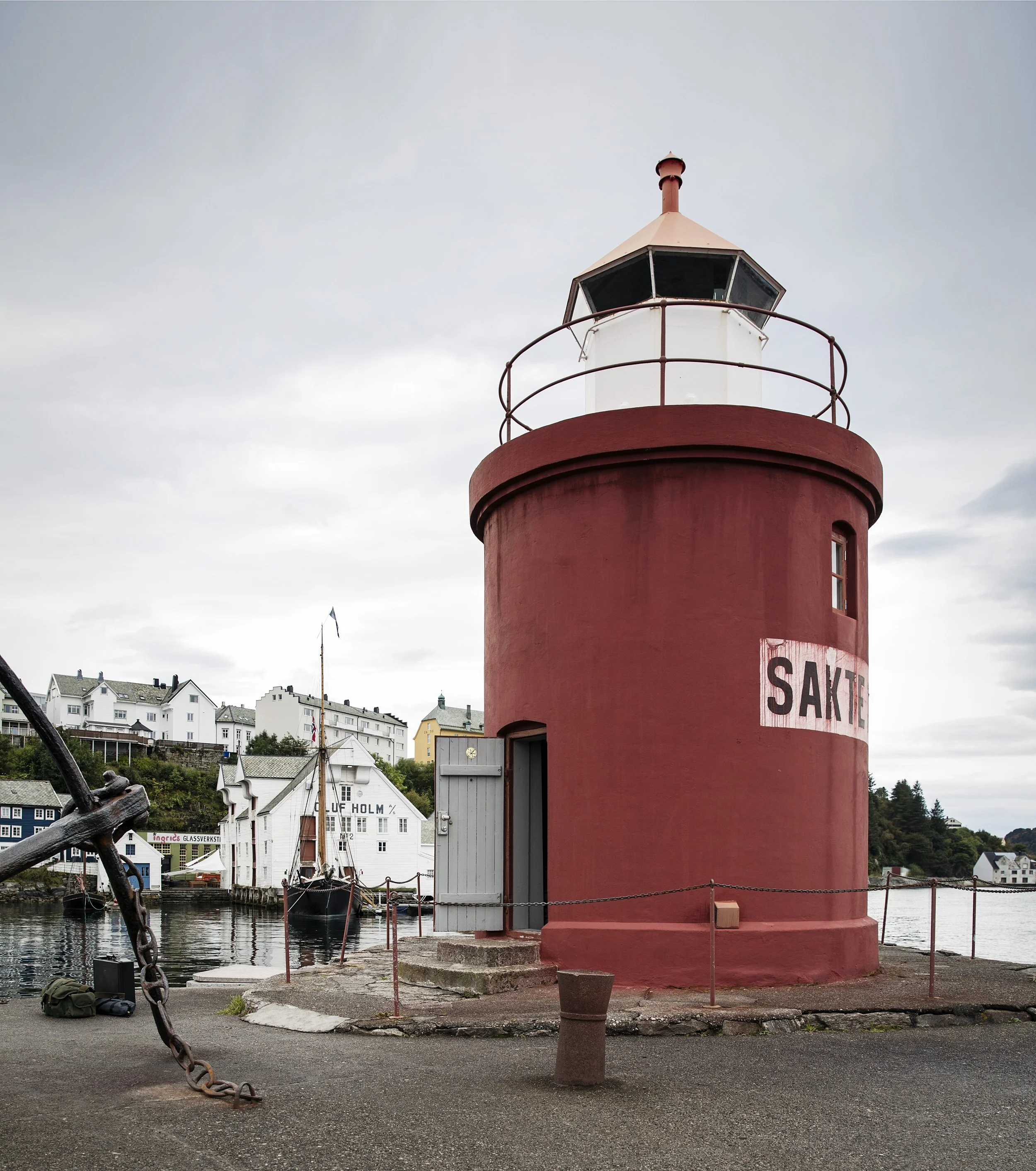 En rød fyrlykter med en liten terrasse omkranset av en rusten metallrekkverk, plassert ved vannet med flere seilbåter og hus i bakgrunnen.