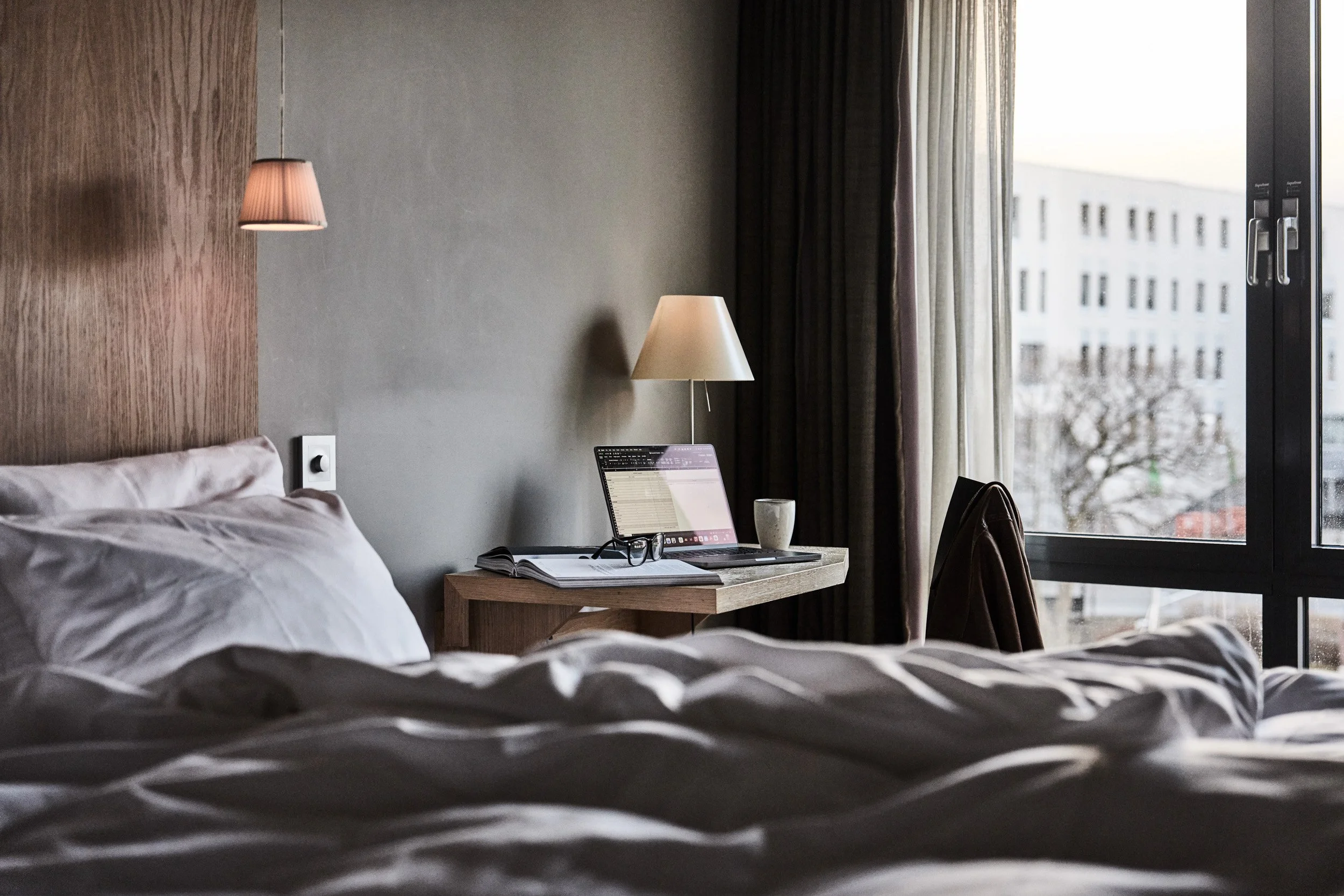 Soverom med seng, nattbord, laptop, bok, kopp, leseelektronikk, lampe og vindu.
