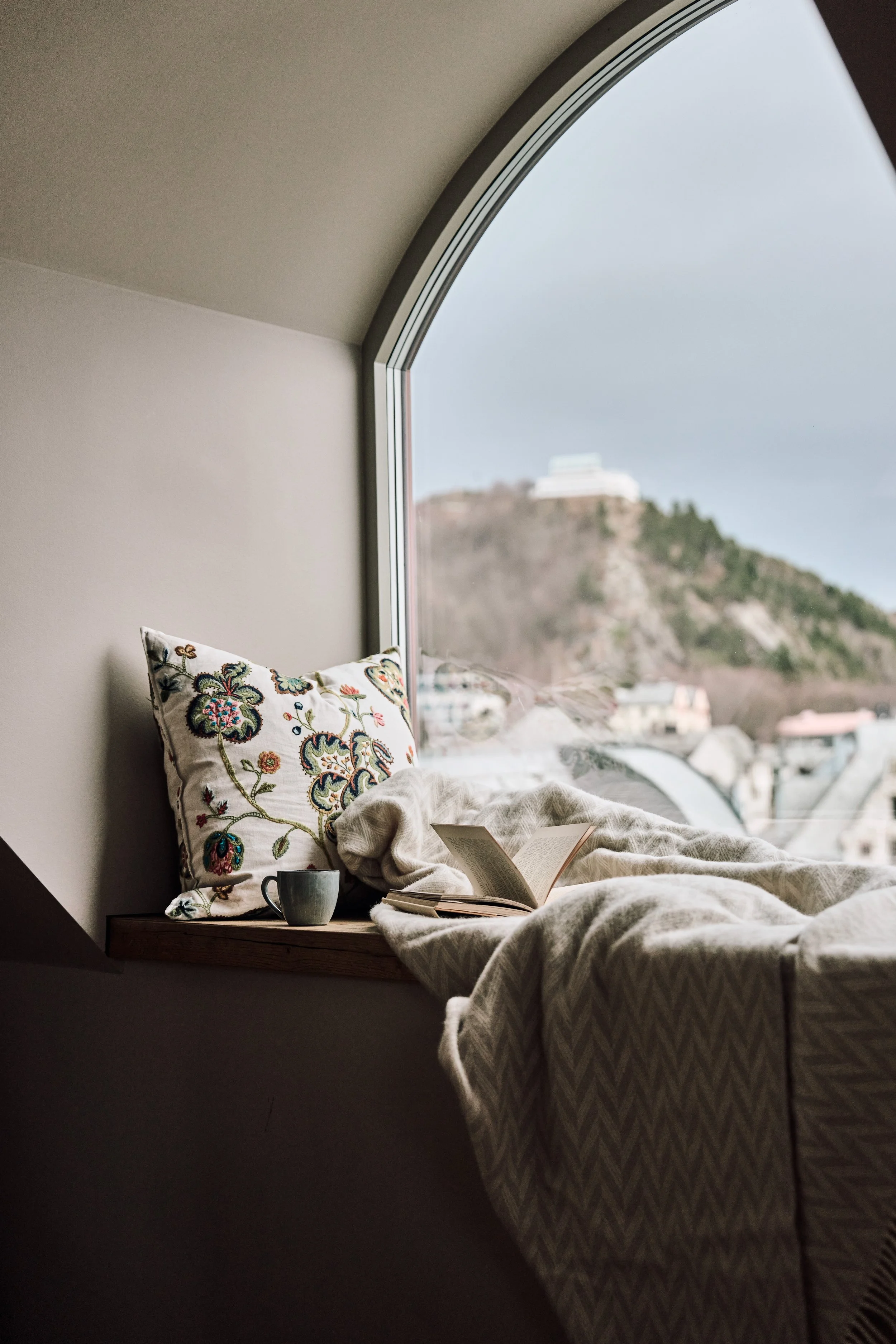 Soverom med en stor vindusvegg som vender ut mot ås med hus på toppen. Sengen er dekket med en hvit dyne og har en dekorativ pute med blomstermønster. På nattbordet står en kopp og en åpen bok.