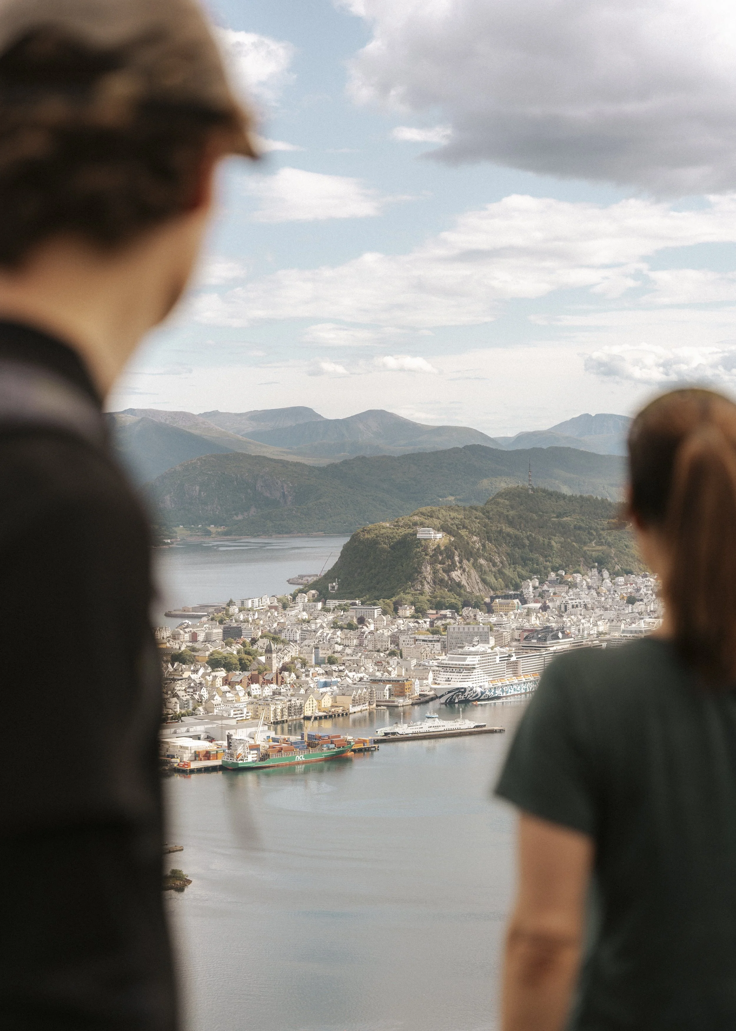 Utsikt over en havn med cruise-skip, bygninger og fjell i bakgrunnen, sett gjennom to ukjente personers skuldre.