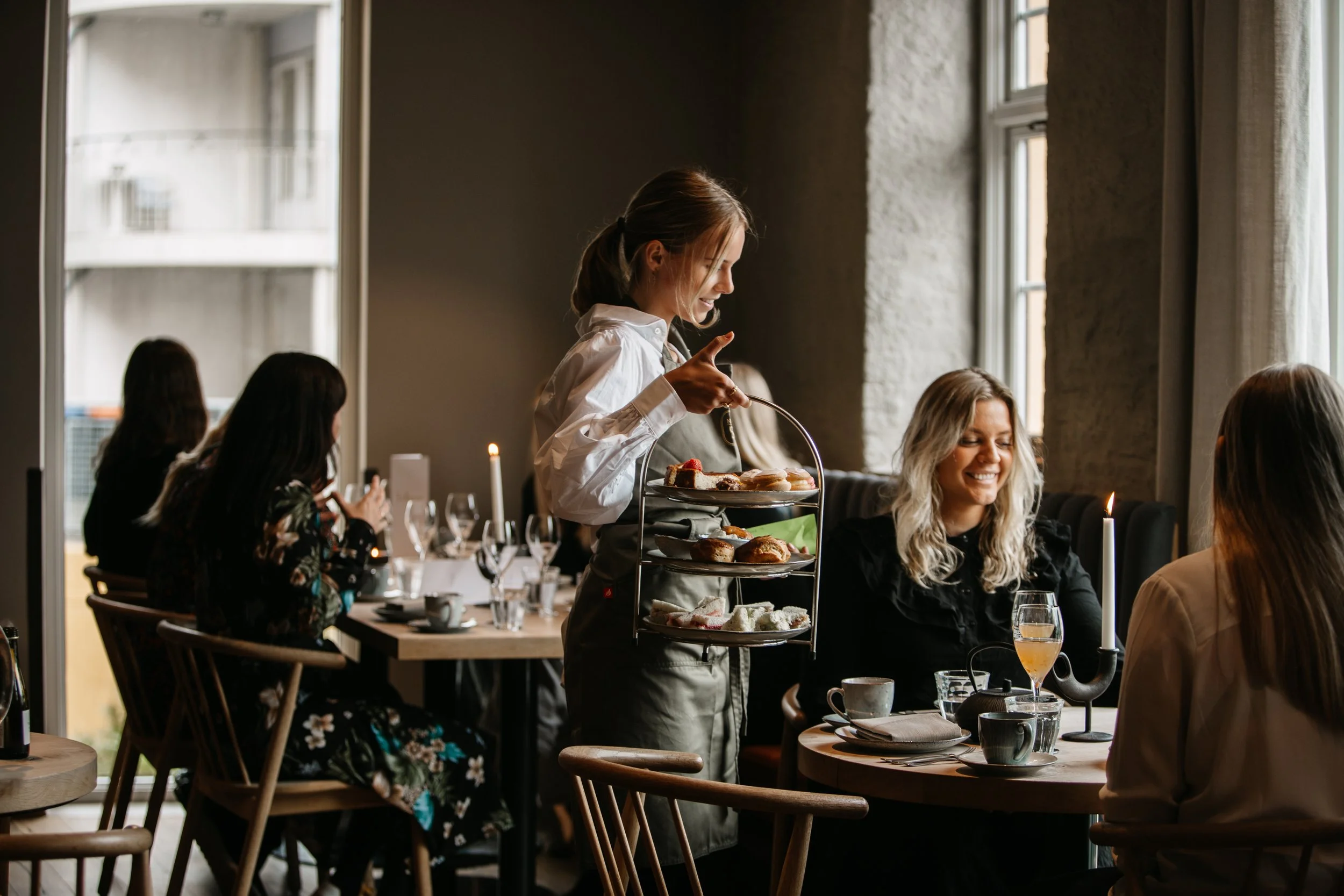 En servitørinne bærer en kakefat med ulike typer kaker til et bord i en restaurant med flere gjester.