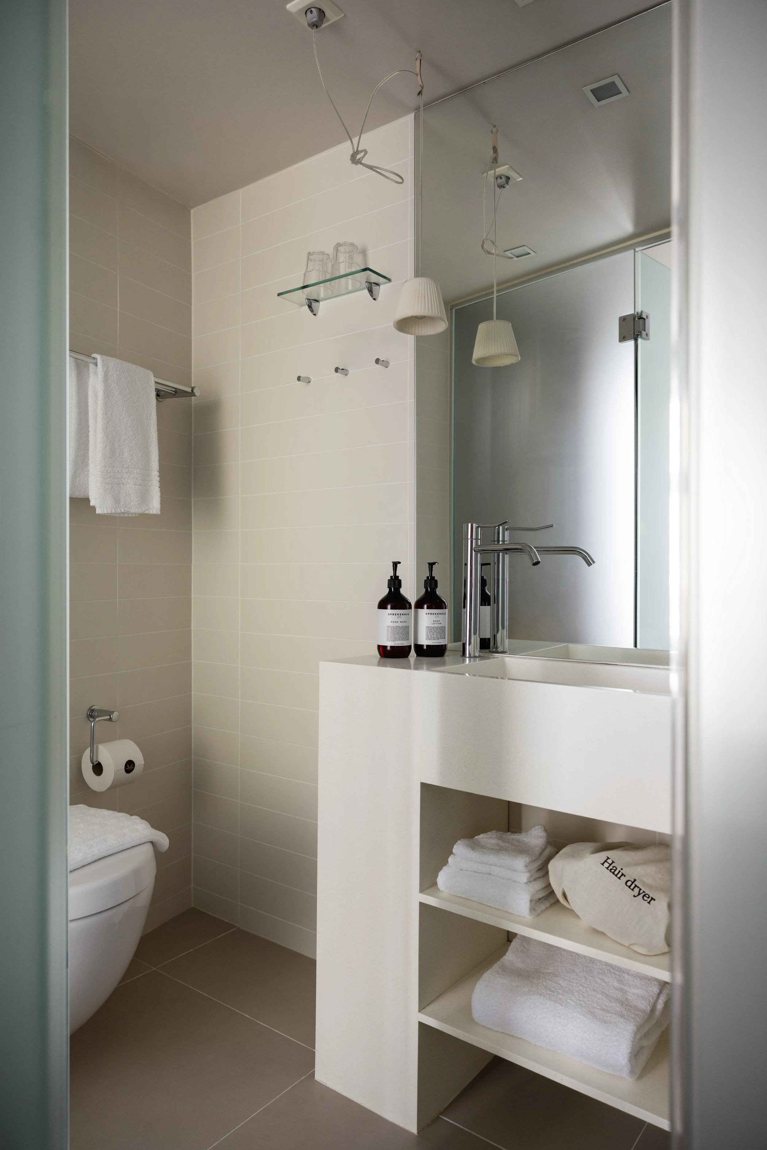 Moderne bad med hvite vegger og mørke gulvfliser. Badet har en hvit servant med en stor speil over, og to brune såpe- eller lotionflasker på vasken. På høyre side er det åpne hvite hyller med håndklær og en pose merket 'Hair dryer'. En halvpart av toalettet er synlig i venstre hjørne, med en hvit håndkle over. På veggen er det en hylle med tre tomme glass og flere kroker uten noe hengende på. To hengende lyslamper og en taklampe lyser opp rommet.