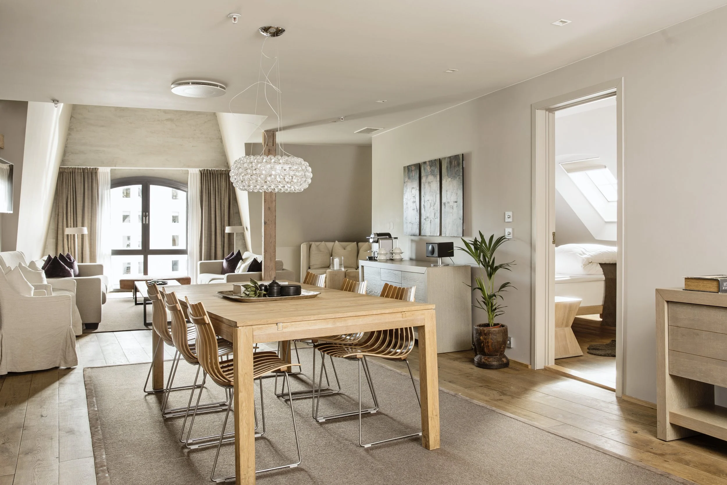 Lys og romslig stue med store vinduer, komfortable sofaer, tre spisebord, kunst på veggen, grønn plante, og et soverom med skråtak i bakgrunnen.