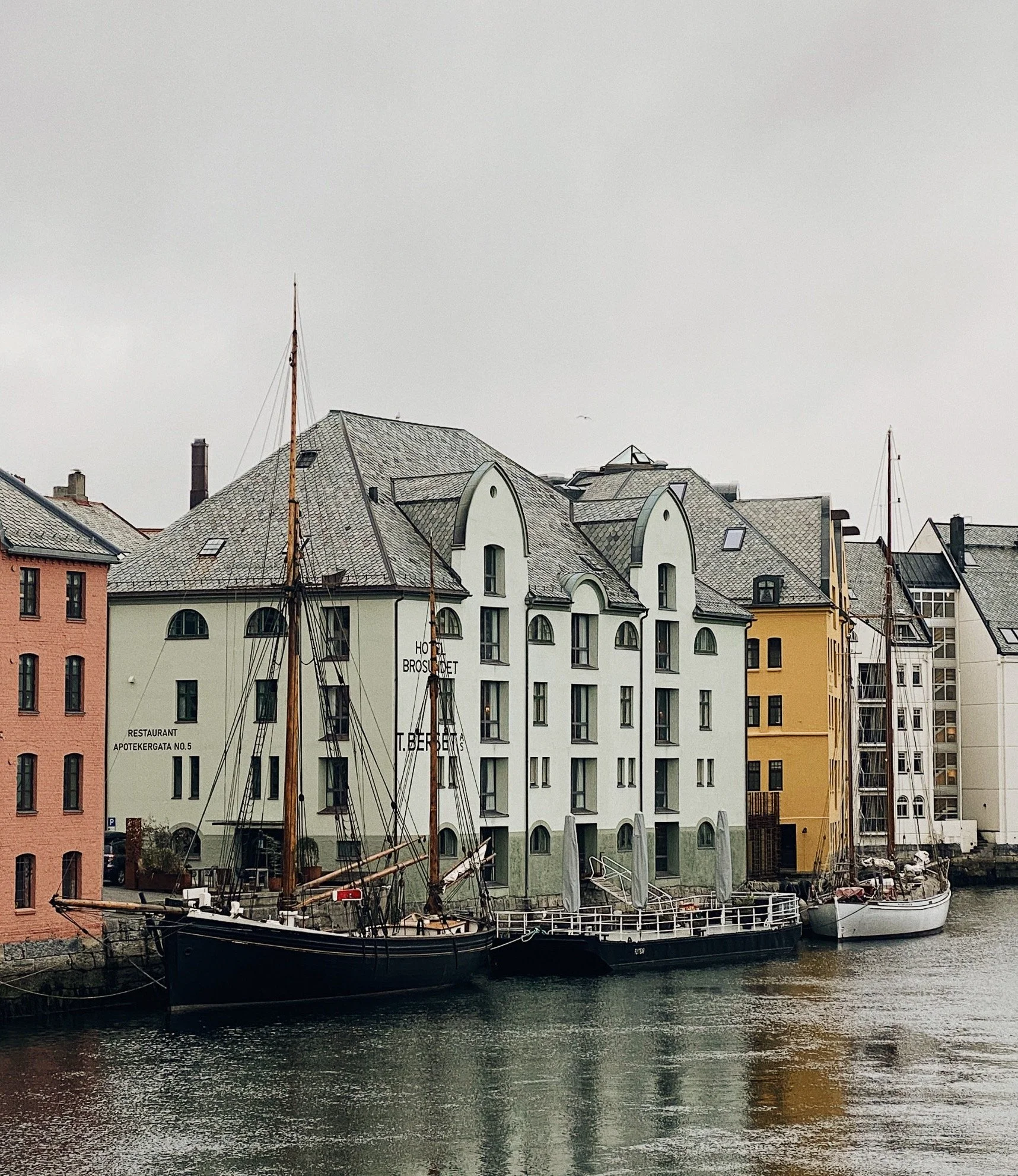 Fabrikker og bygninger langs vannkanten med båter i vannet foran. Over er det en grå himmel.