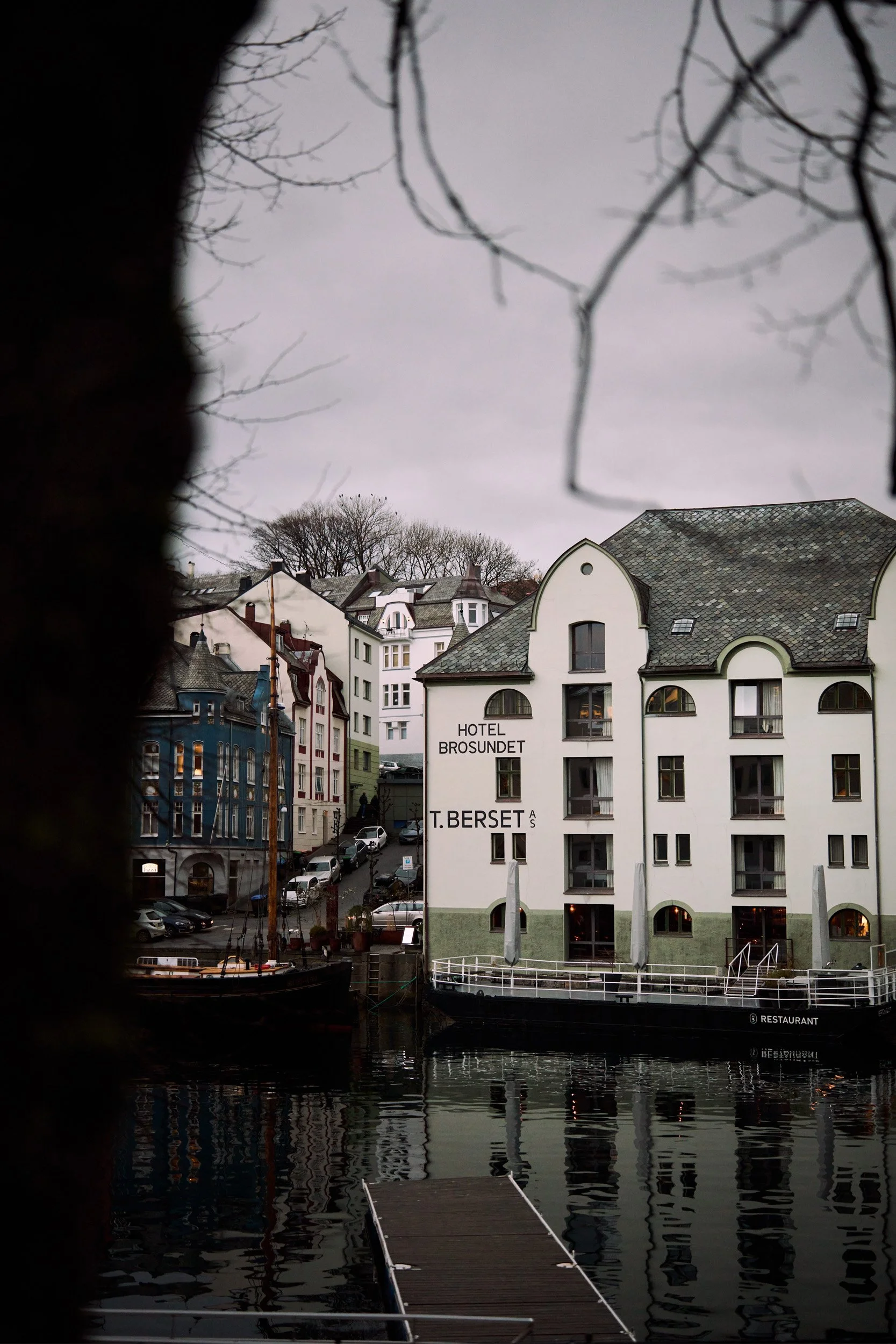 Utsikt over by med flere bygninger, en havn med en brygge, og trær med uten blader. Det er overskyet vær.