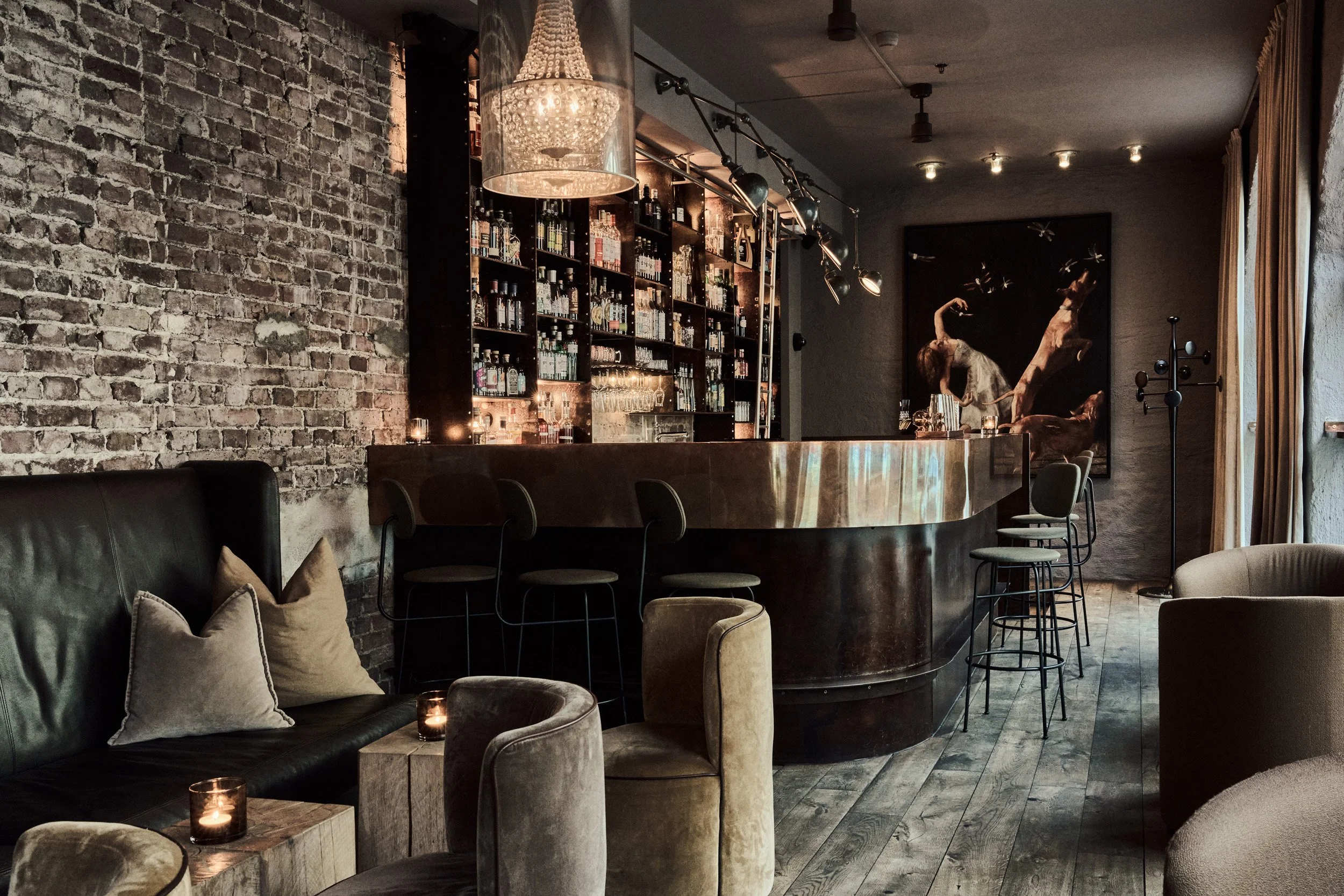 Elegant bar med brune og grå møbler, en rustikk bakvegg av murstein, en stor kunstinstallasjon med en kvinne og flyvende insekter, samt diverse flasker og glass på hyller.