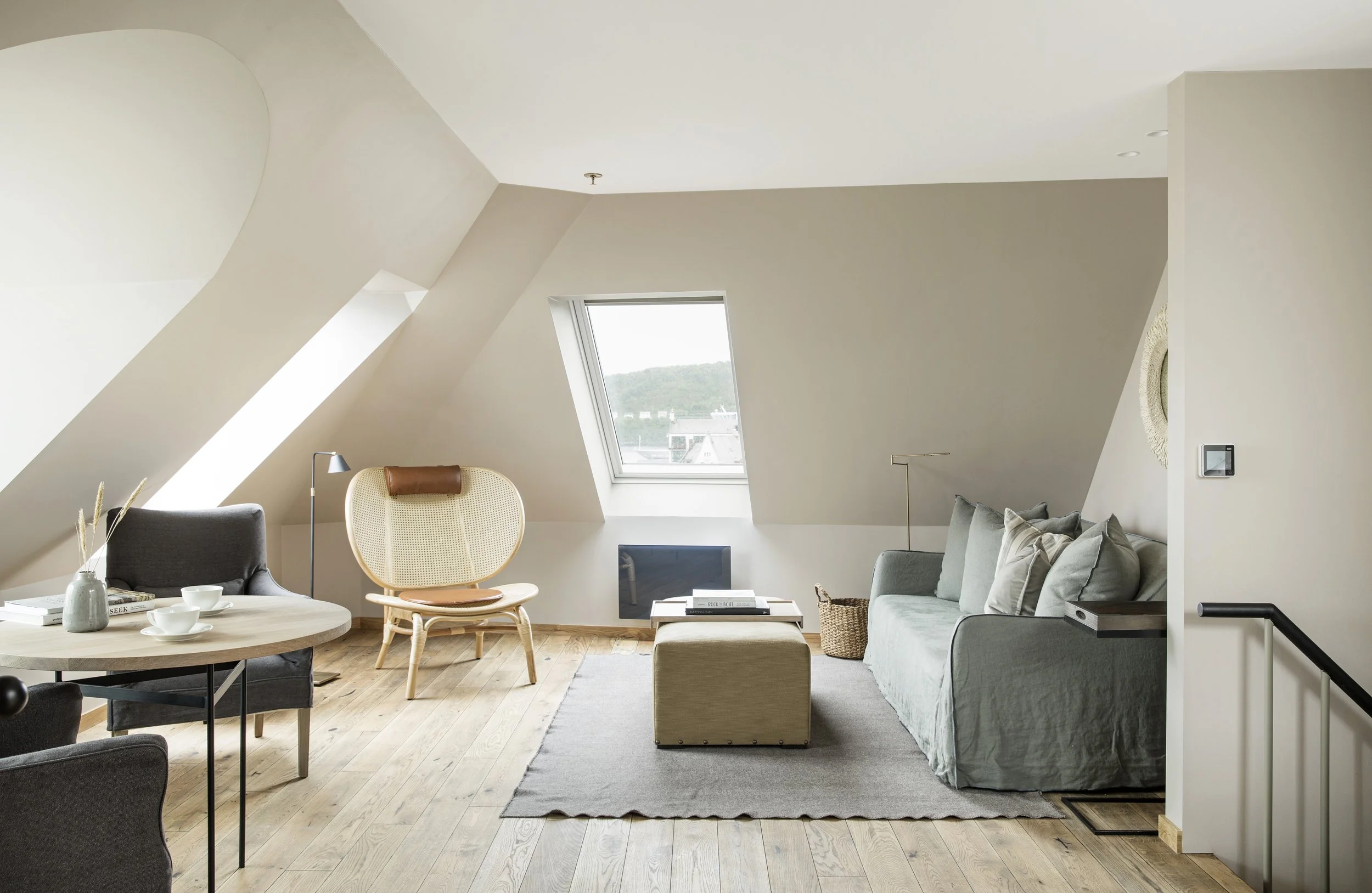Et lyst og moderne stueområde med skråtak, et vindu med utsikt over landskap, en grå sofa med puter, en beige lenestol, et rundt trebord med kopper og bøker, en gul stol og ulike dekorative elementer.