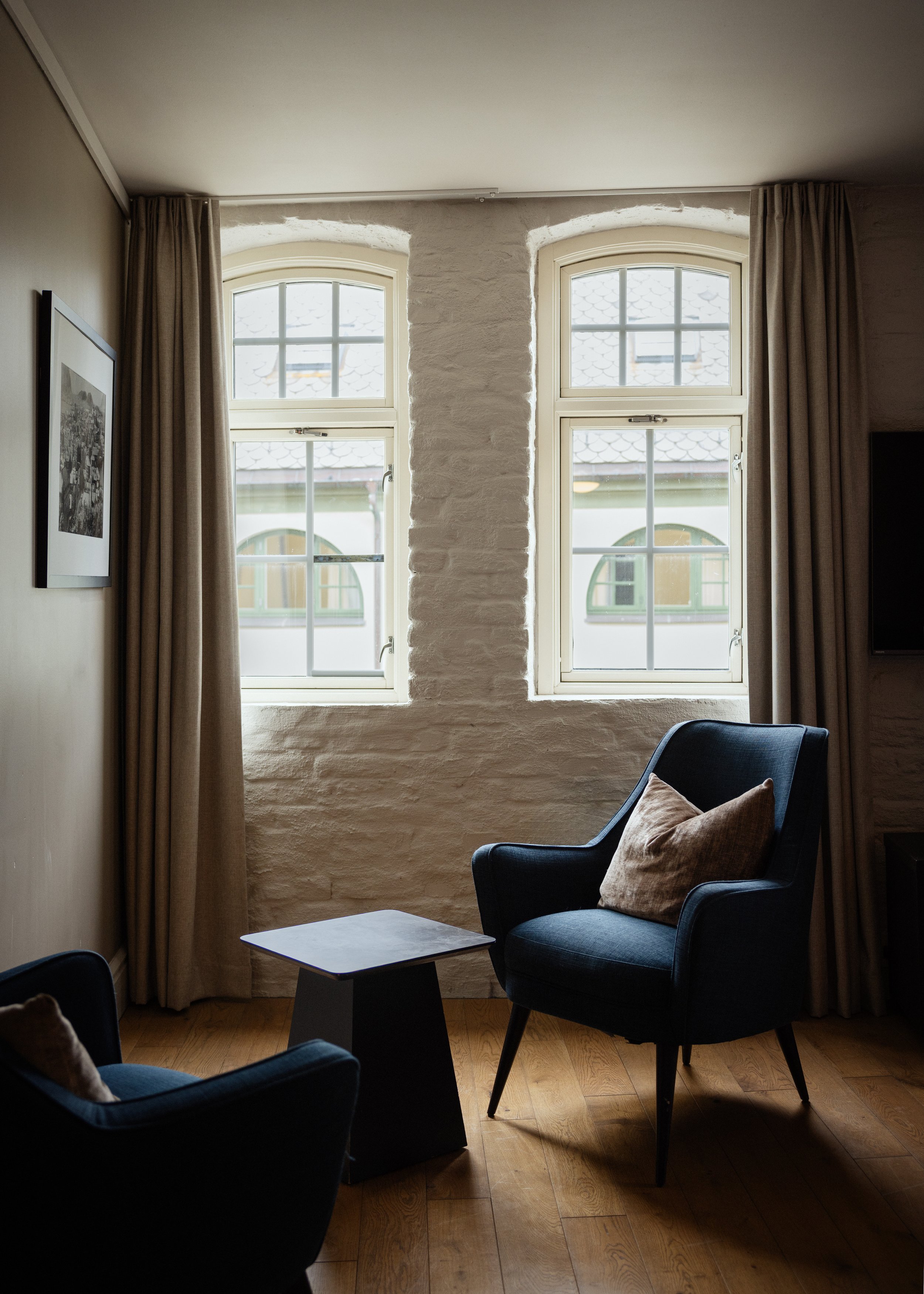 Innendørs bilde av to vinduer med hvite rammer, beige gardiner, en svart armstol med puter, og et lite bord på en tremålt gulv
