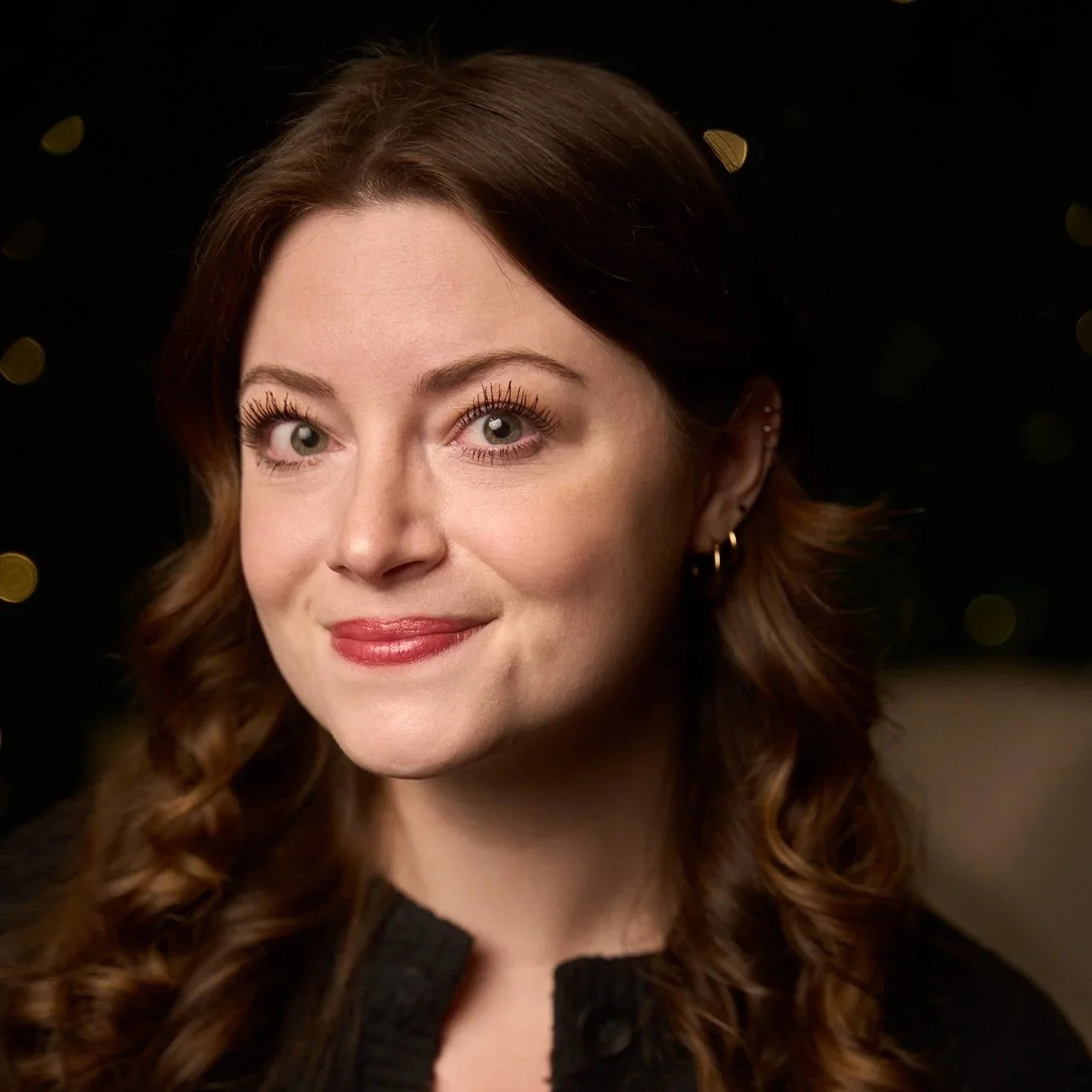 Portrait of a woman with wavy brown hair and hoop earrings smiling, black background with soft light highlighting her face