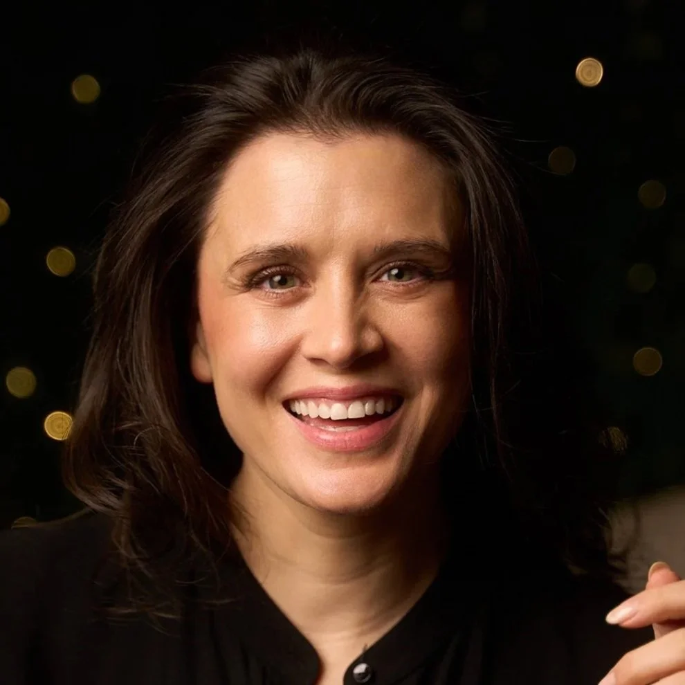 A woman with dark brown hair smiling at the camera against a dark background with blurred lights.
