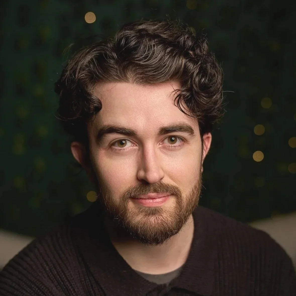 Portrait of a young man with dark, curly hair and a beard, wearing a dark sweater, smiling softly against a blurred dark green background with bokeh lights.