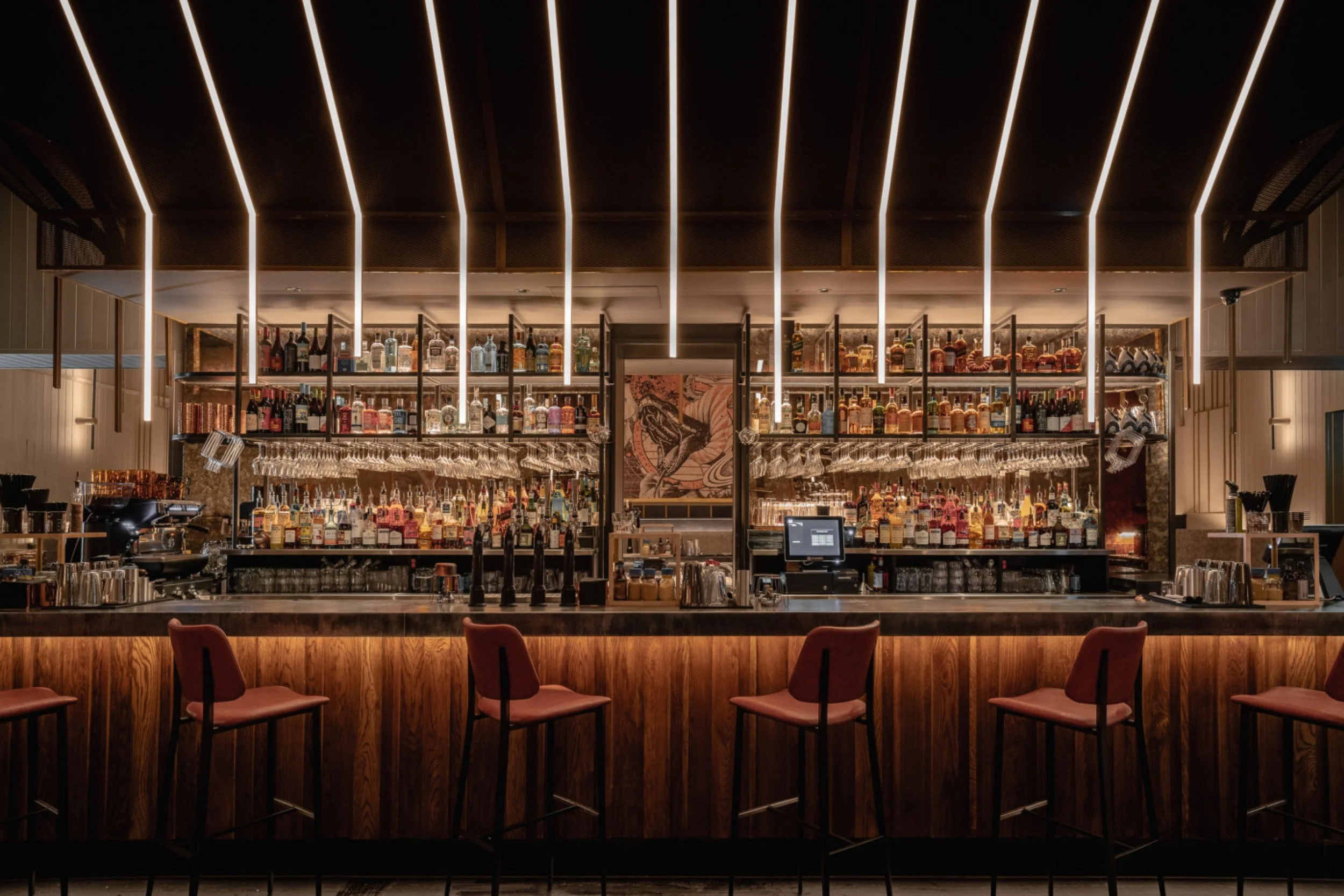 Modern bar with illuminated ceiling lights, shelves filled with various liquor bottles, garnished with wine glasses, and cozy seating in the foreground.
