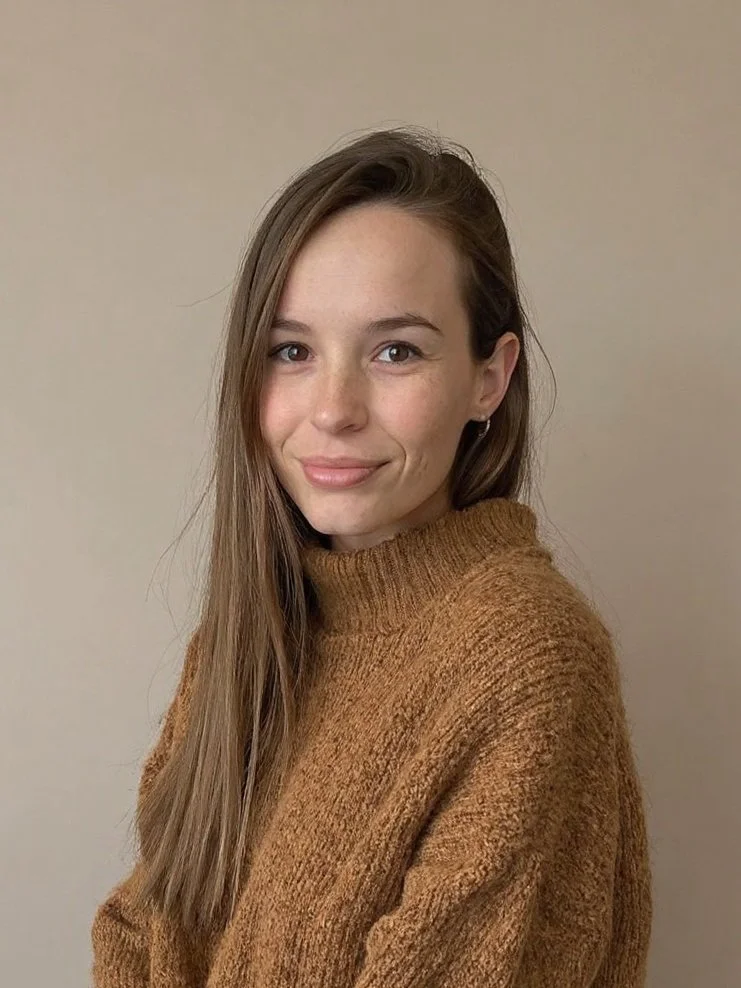 A young woman with long brown hair, smiling softly, wearing a brown knit sweater, standing against a plain beige wall.