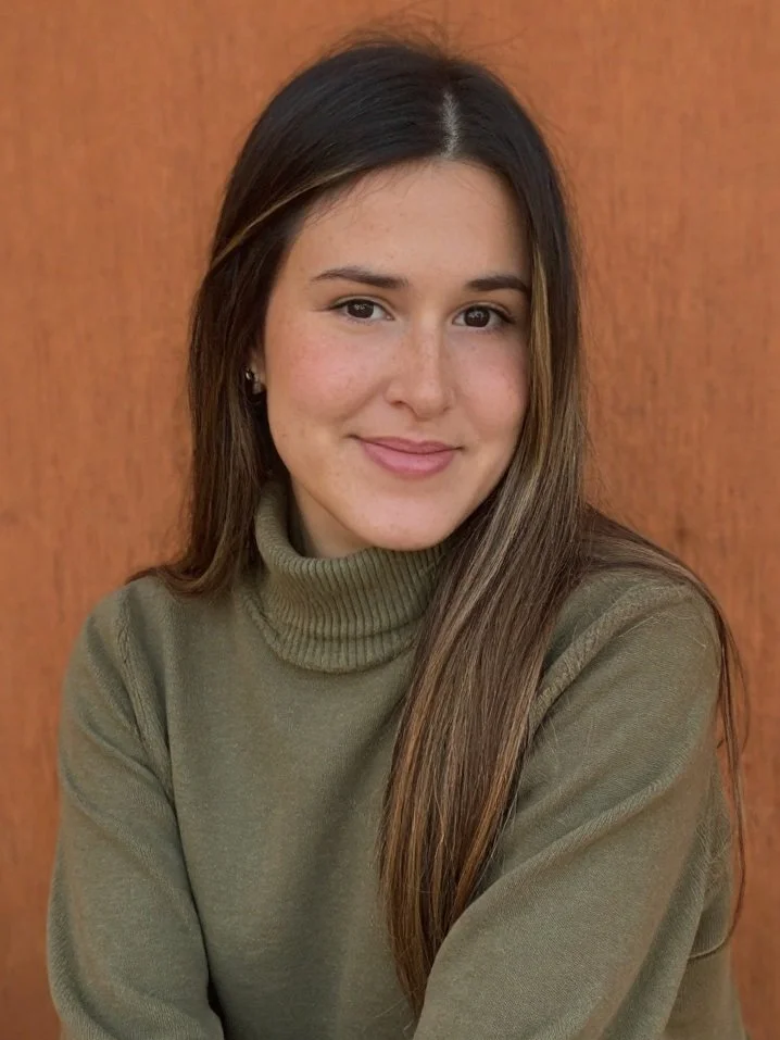 A young woman with long brown hair, wearing earrings and a green turtleneck sweater, smiling softly in front of a wooden background.